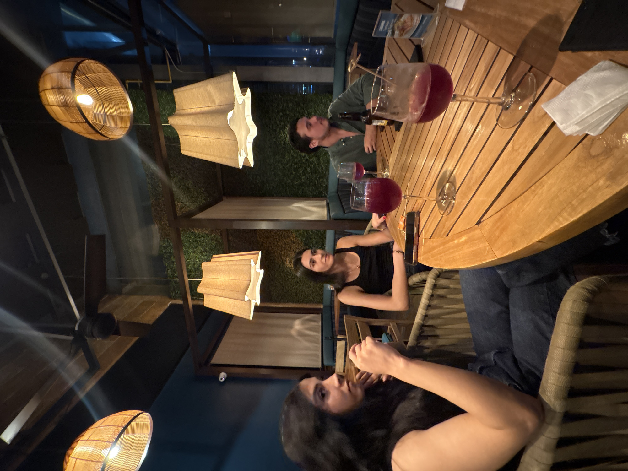 At a bar or restaurant in Puebla, Mexico, a group of four people are sitting around a wooden table drinking red wine. A man sits to the left with a green shirt and a bottle of beer. Two women, one in a black tank top and the other with long dark hair, sit closer to the viewer. The table has a stack of white napkins, and there are two glasses of red wine. The scene is lit by decorative hanging lamps and ceiling fan.