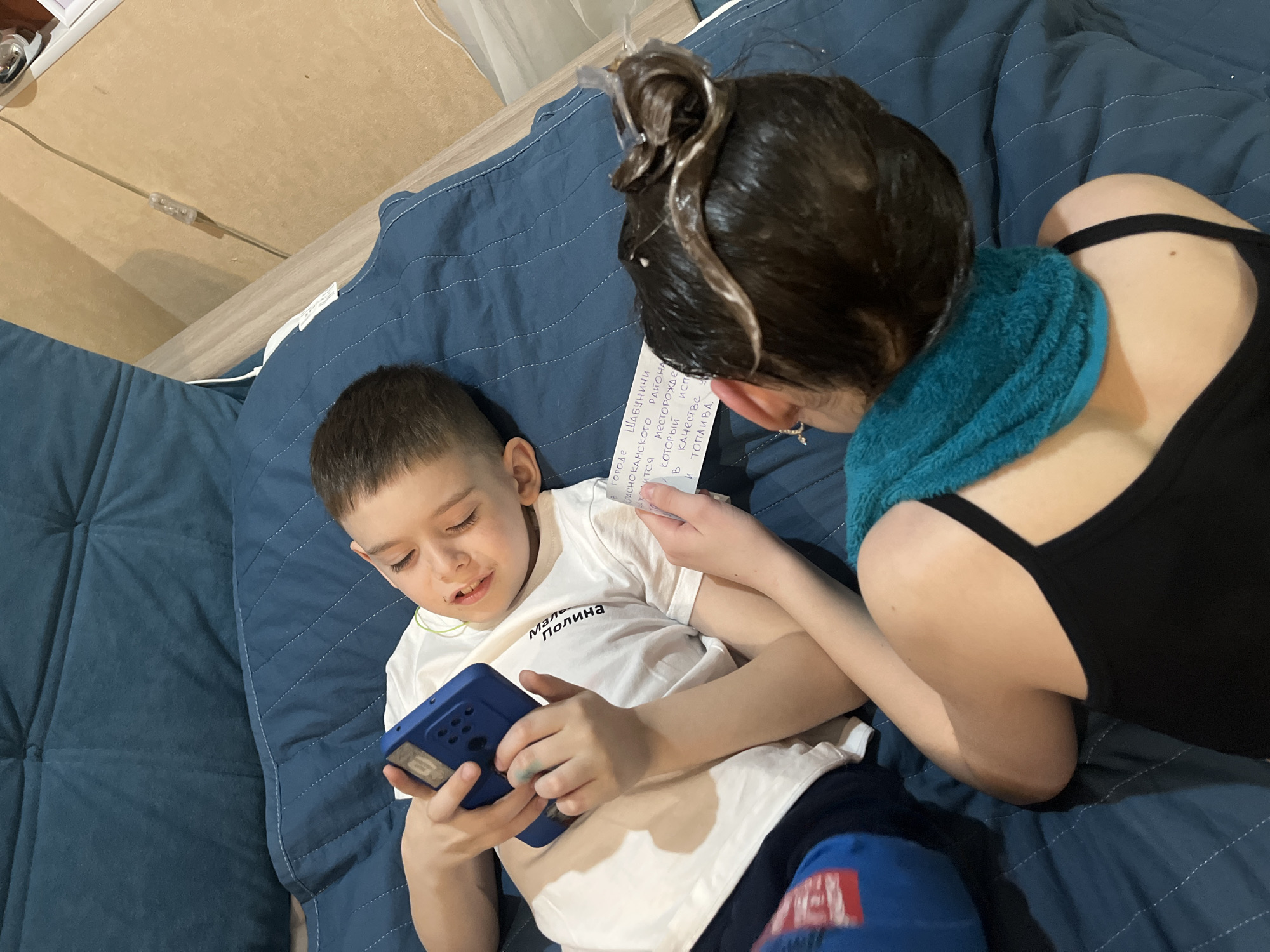 A boy, presumably lying on a bed covered with a blue quilted blanket, is engrossed in a blue device with a screen, likely a phone or small tablet. He is wearing a white t-shirt with the word "Maль Полина" written on it, with blue pants.
Next to him sits a woman, partially visible, wearing a black tank top and having a blue towel around her neck. Her hair appears to be covered in hair dye. She holds a piece of paper with Russian text, seemingly reading it to the boy. The paper contains the text: "В городе ШАБУНИЧИ РАСНО КАМСКОГО РАЙОНА ... КОТОРЫЙ ИСПЕ ... в качестве и топлива."
The scene seems to be indoors, possibly in a bedroom. Part of the bed frame or side table is visible on the left.