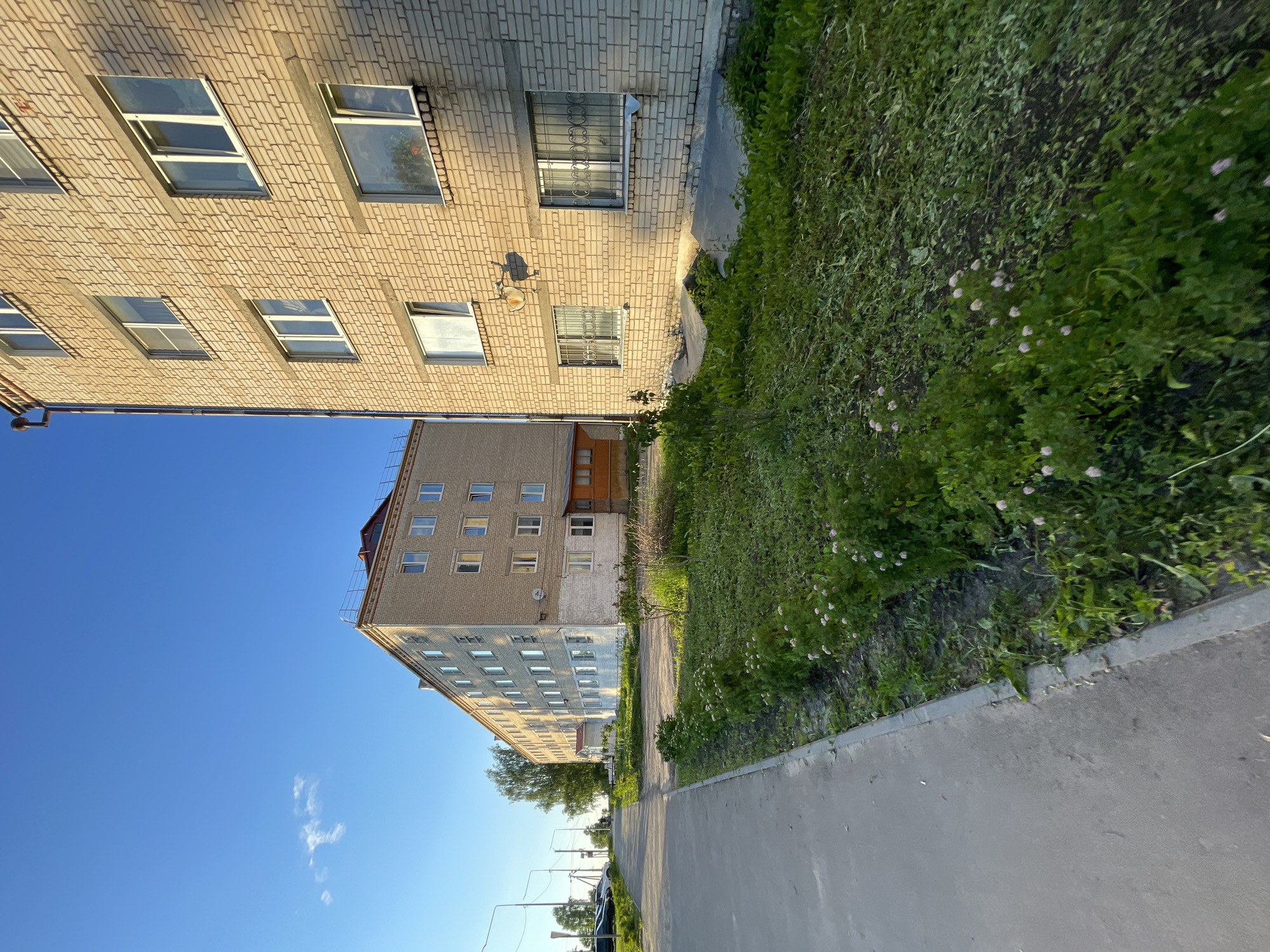 In the village of Krasnye Baki, Russia, a residential street is depicted under a clear blue sky with scattered white clouds. A multi-story brick building, possibly an apartment complex, dominates the right side of the frame, its light-colored bricks illuminated by the setting sun. The building has numerous windows, some with protective bars. In the foreground, a concrete sidewalk runs along the left edge of the image, bordered by a strip of green grass and small pink wildflowers. To the left of the sidewalk, cars are parked along the street. Another, older building with a similar multi-story structure and brick facade is visible in the distance on the left, receding down the street. The overall scene suggests a quiet, residential area during the late afternoon or early evening.