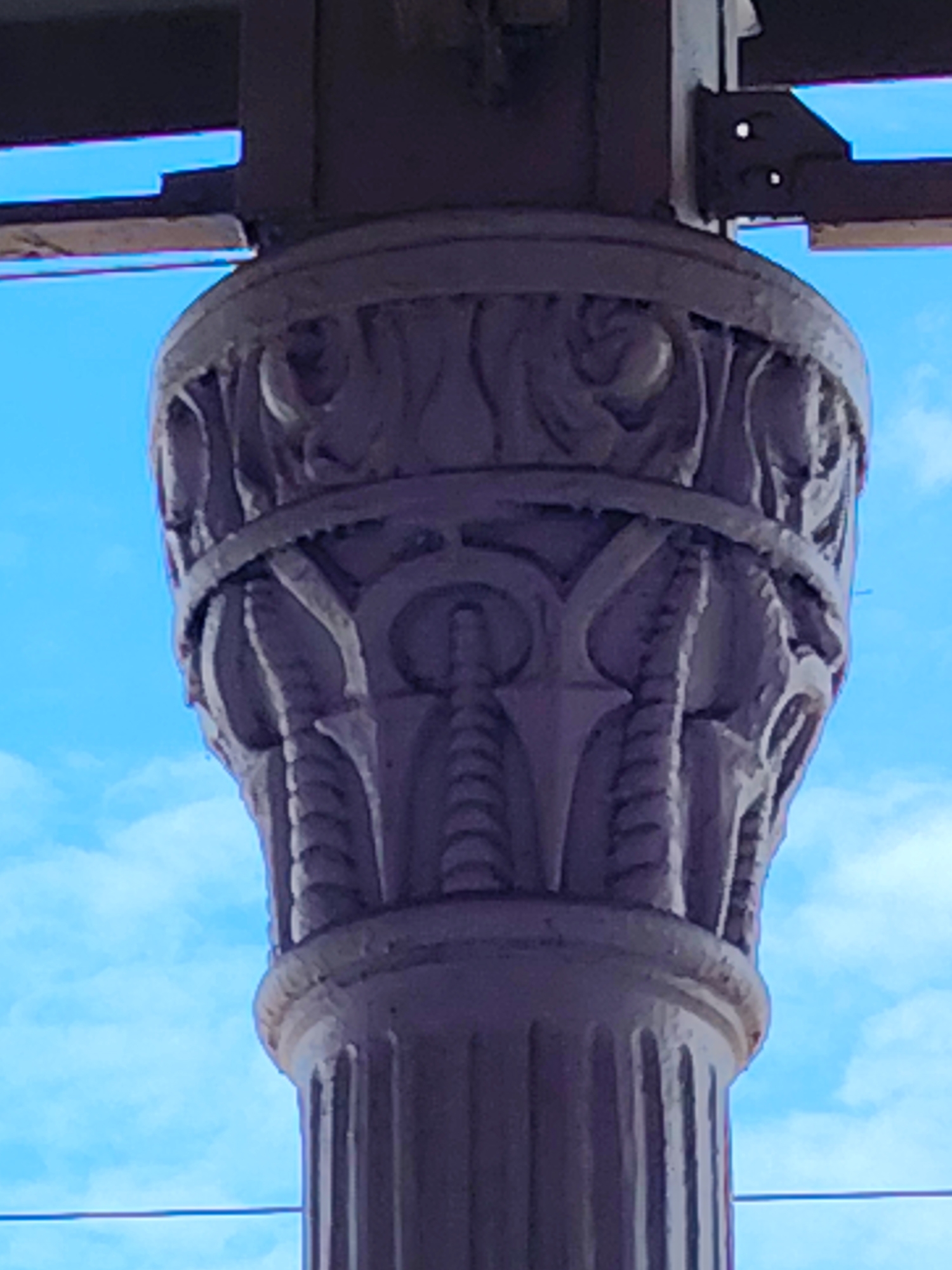 The image shows a close-up, low-angle view of a decorative column, likely made of stone or concrete, against a bright blue sky with scattered clouds. The column features ornate carvings. The top section displays a band of swirling, leaf-like motifs, followed by a ribbed section with a circular, geometric design in the center. Below this, the column is fluted. In the upper background, a dark structural element, possibly part of a bridge or overhead structure, is visible, along with a thin horizontal wire. The image is cropped tightly on the column, with no people or other contextual elements of the city of Klin, Russia visible.