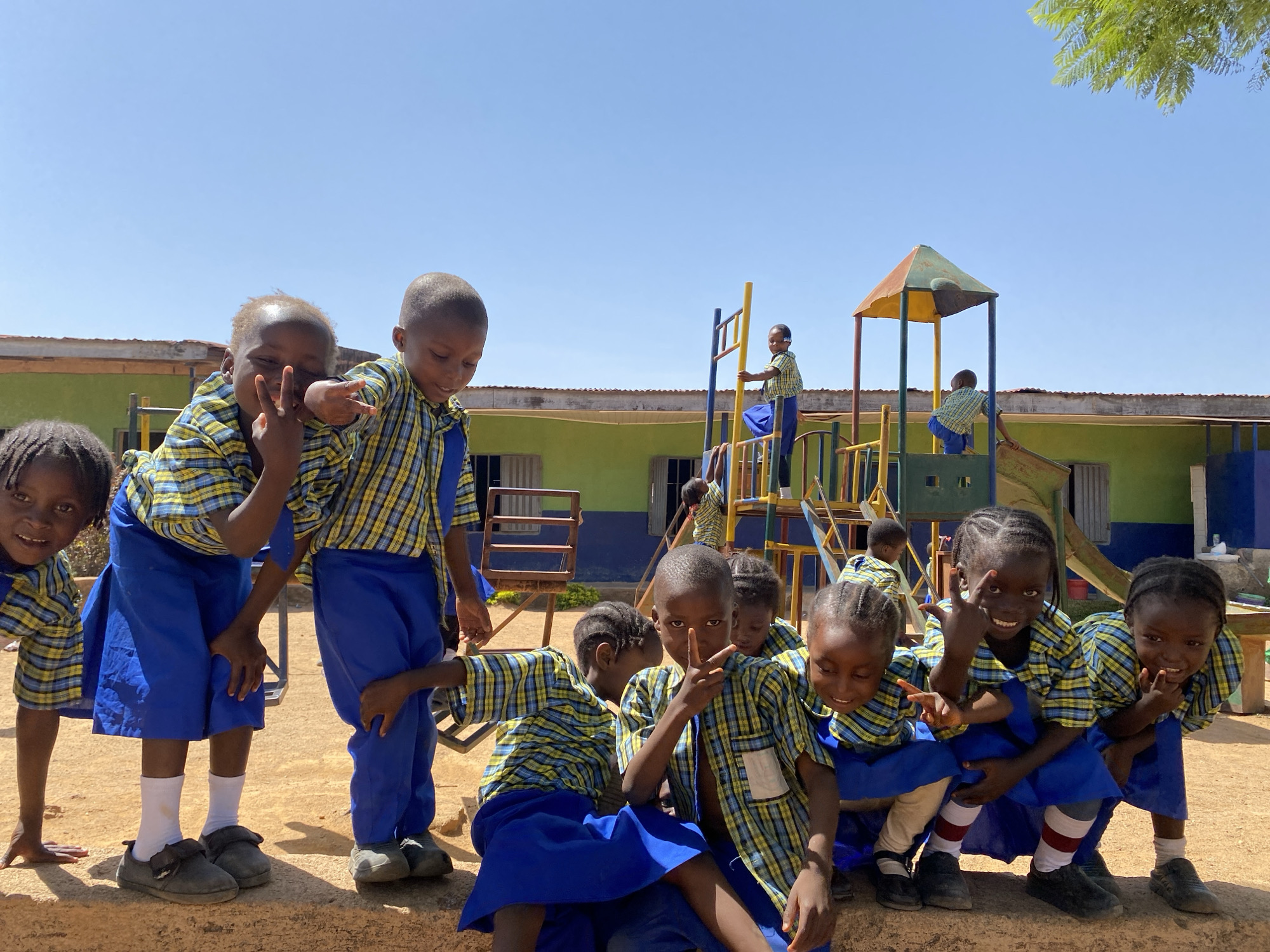 A group of children in uniforms are on a playground in Gura Top, Nigeria. The uniforms consist of blue skirts or shorts and plaid shirts with blue, yellow, and black colors. The children are standing or crouching near the front, some making peace signs with their fingers. Other children are on the playground equipment behind them, including a slide and climbing structure. The building behind the playground is painted green and has a tan-colored roof. The sky is clear blue. The environment appears sunny and dry.