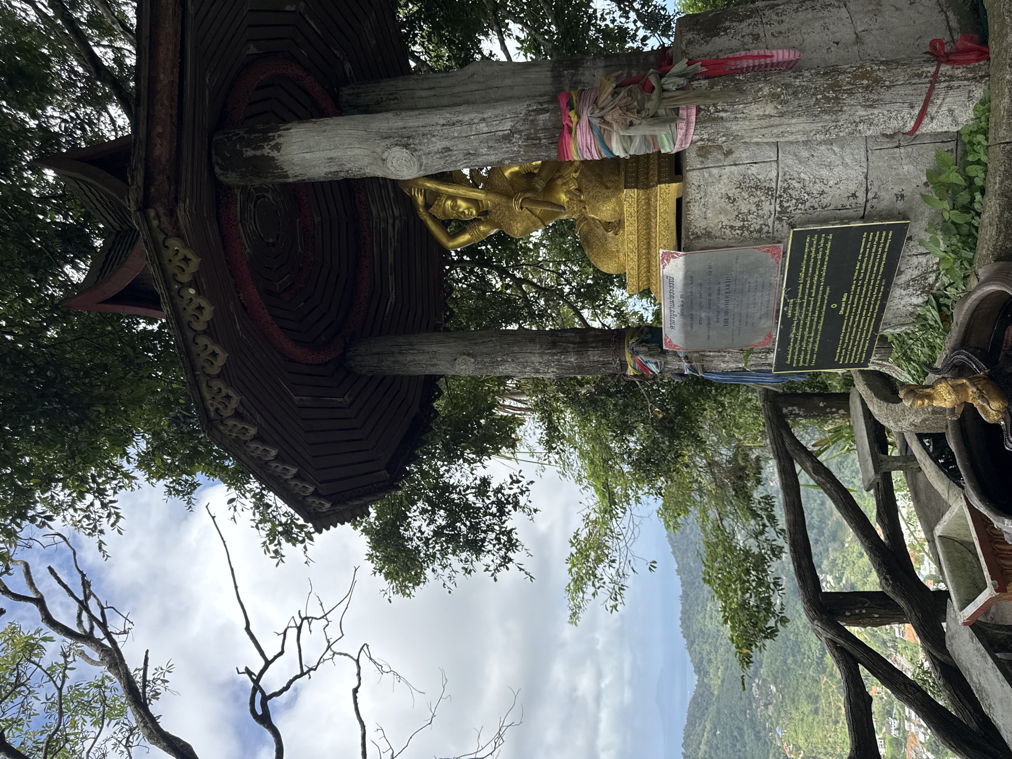 The image depicts a shrine or religious structure in Karon, Thailand. At the center is a golden statue, possibly representing a deity, placed on a platform supported by wooden pillars. The platform is decorated with colorful ribbons. Two plaques with Thai and English inscriptions sit to the right of the statue, bearing information about the Mother Earth goddess Dharani Mata. The structure is partly covered by a wooden roof adorned with traditional designs. Trees and greenery surround the shrine, with a view of the mountainous landscape and cloudy sky visible in the background. Part of a wooden railing can be seen on the right, and bare tree branches are visible in the lower-left corner.