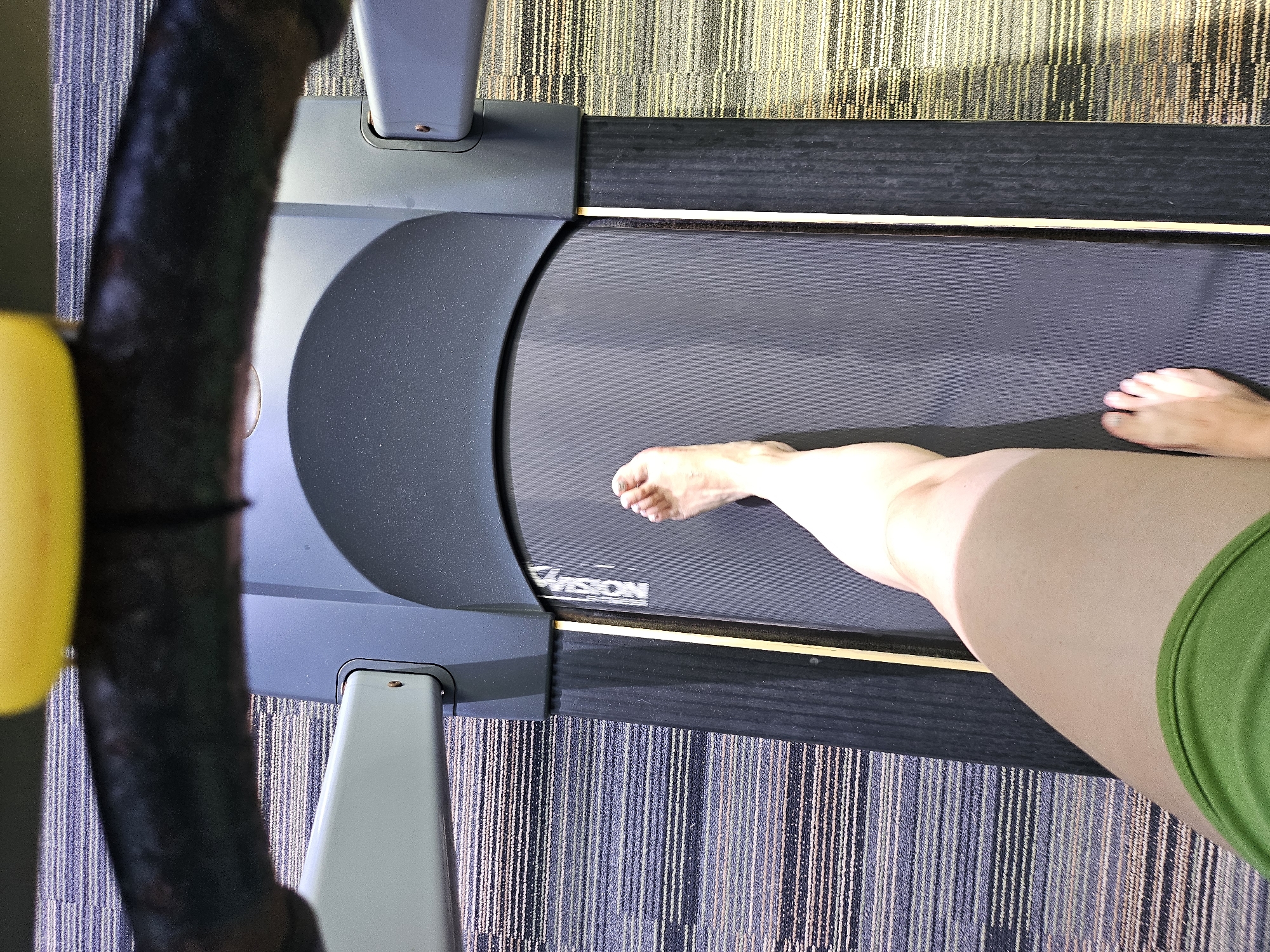 An overhead, close-up shot shows a person's bare left foot stepping onto a treadmill belt, mid-stride. The treadmill is gray with a black belt and a yellow handlebar is visible at the top of the frame. The person is wearing a green garment. The surrounding area appears to be a gym, with striped carpeting visible. The location is specified as Kathu, Thailand.