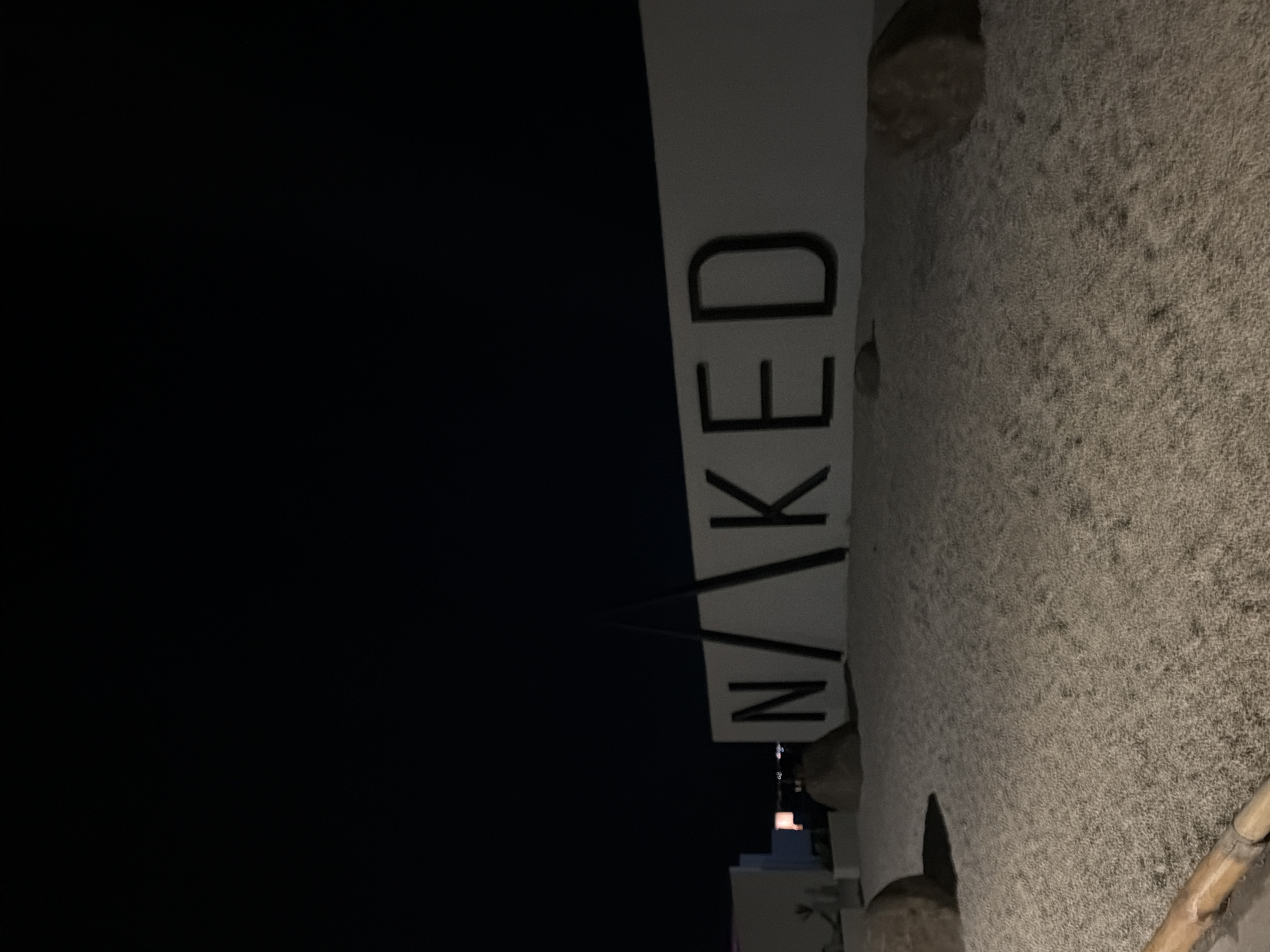 A sign with the letters "N/AKED" is displayed on a white wall at night. The letters are large and black. The ground in front of the sign is covered in small white pebbles. Several large rocks are scattered on the ground. A tall, thin black structure is positioned in front of the sign, obscuring part of the letters. The sky is dark. The location is the Naked Beach in Burgas, Bulgaria.