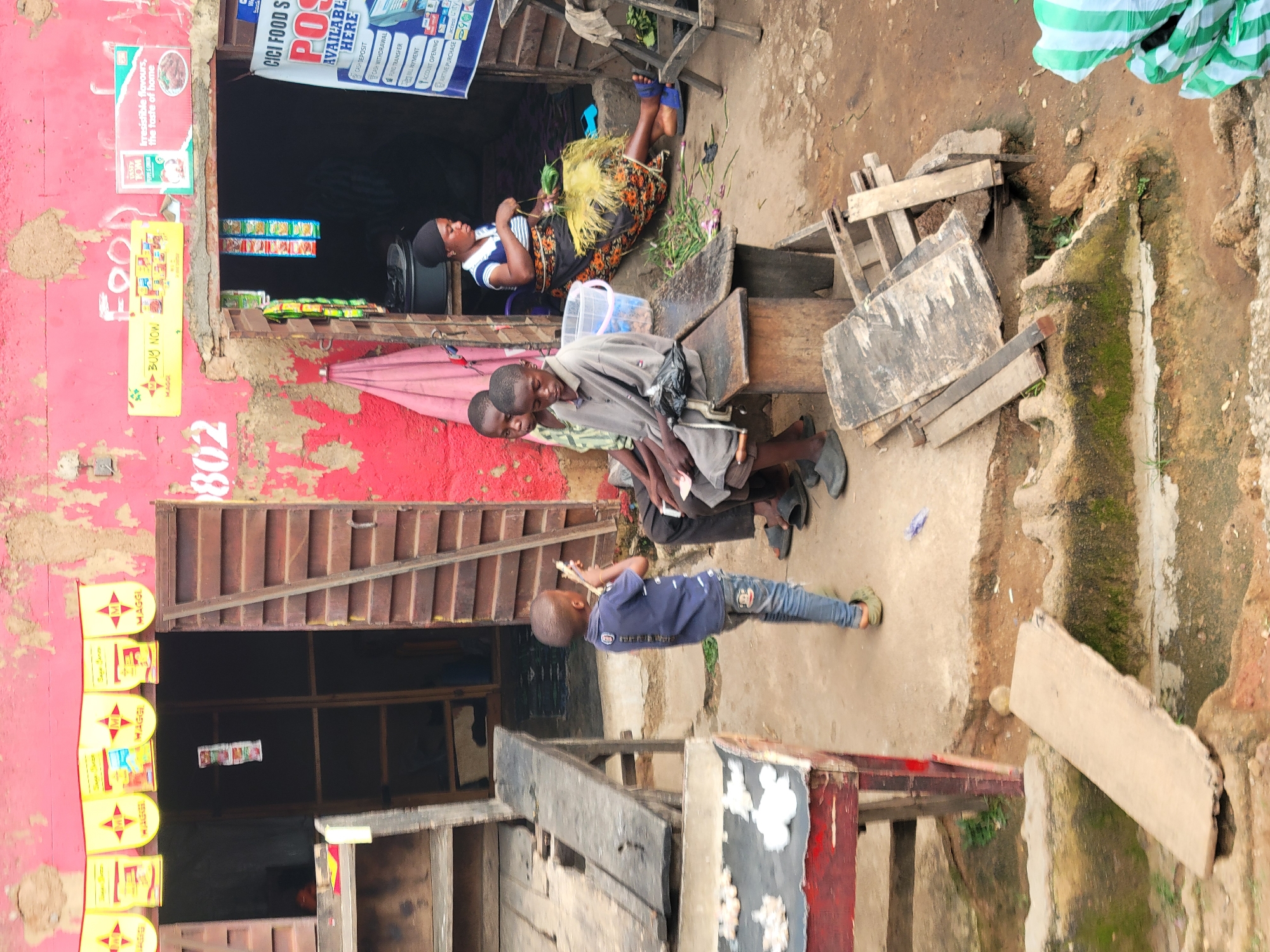 The image depicts a scene in Jos, Nigeria, likely a market or a roadside stall. In the foreground, a young boy in a dark blue t-shirt and jeans stands facing away from the camera, holding a small, thin object. To his left, a red and black table with white items is partially visible. Further back, three young boys are seated on a wooden bench. The boy in the center wears a grey t-shirt and appears to be holding something in his lap. The boy to his right is wearing a light green shirt and is looking towards the boy in the grey shirt.
Behind the boys, a woman with a dark headscarf and a patterned skirt is seated. She is wearing a blue and white striped top and is holding a bunch of green vegetables and some yellow material. She is engaged in preparing the vegetables. The environment appears to be an outdoor market setting with weathered buildings and wooden structures. A pink umbrella is leaning against a red wall. Signs are visible on the building, including one that reads "BUY NOW" and another with "CICI FOOD STORES". The ground is dirt and concrete. In the far right of the image, a large green and white striped bag is visible.
