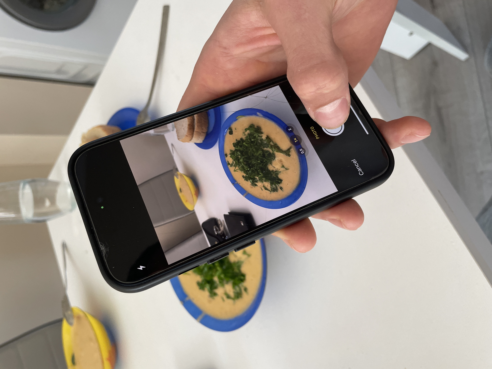 A person is holding a black smartphone to take a photo of a bowl of soup on a white table. The soup is yellow in color and garnished with green herbs, served in a blue-rimmed bowl. A few slices of bread are on a plate next to the soup. A fork is also visible on the table. The person's hand is partially visible as they hold the phone. The background includes a washing machine and a white piece of furniture. There is no indication of this scene being specifically located in the city of Plovdiv, Bulgaria.