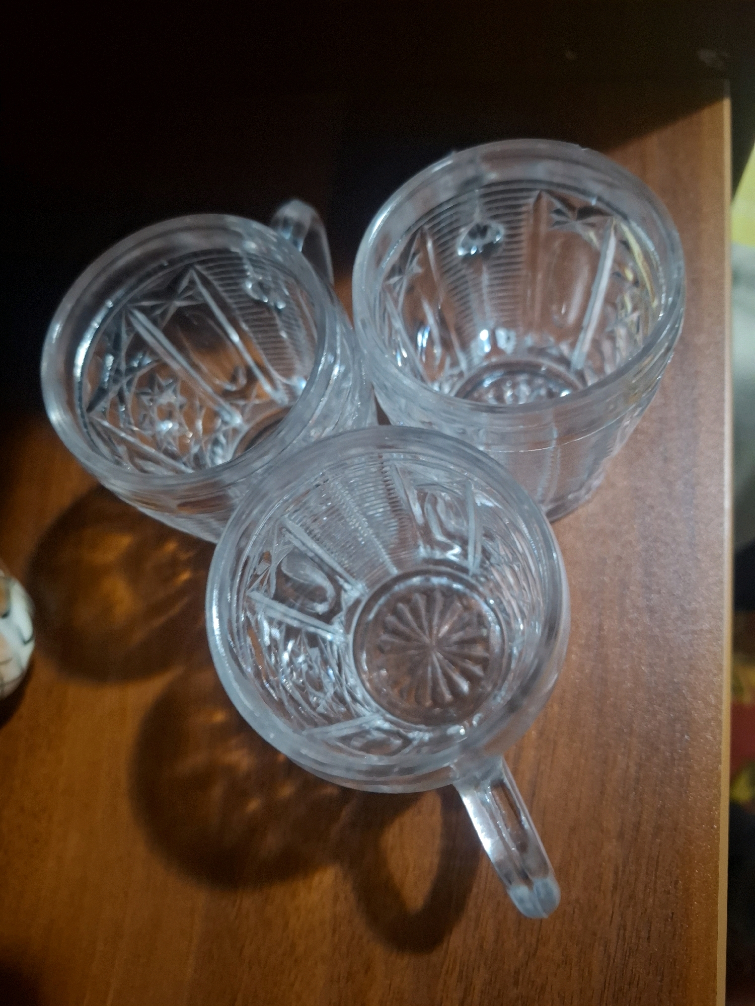 Three clear glass cups with etched patterns are arranged on a wooden surface. The cups are of different sizes and appear to be vintage. One cup is larger and positioned towards the top right, while two smaller cups are positioned in front of it, forming a triangular arrangement. The cups have handles. The wooden surface has a warm, brown tone and a visible grain. The background is dark and out of focus. The lighting suggests an indoor environment, possibly a home setting. The image was taken in Almaty, Kazakhstan.