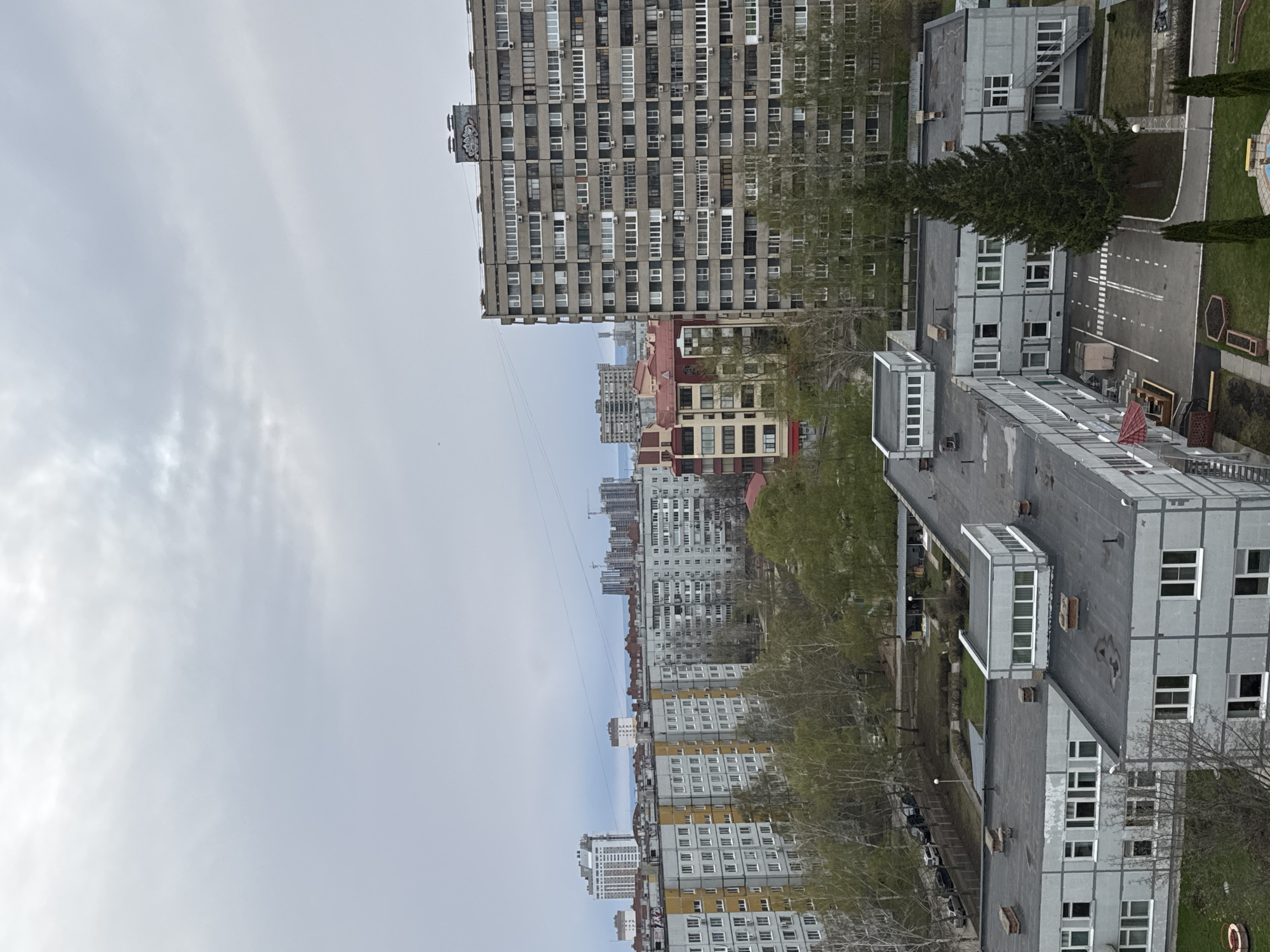 The image displays an urban landscape, likely from a high vantage point, dominated by numerous multi-story residential and utilitarian buildings under an overcast sky. The overall impression is one of a dense, potentially post-Soviet era, development.
The subjects include a variety of buildings. In the foreground and mid-ground, there are several large, blocky apartment buildings, predominantly gray, with repetitive window patterns and numerous balconies. One particularly tall, grey building with many small windows and air conditioning units is prominent on the left side of the frame. On its upper edge, a small, grey structure features a stylized logo or graffiti in white. To the right and closer, several lower-rise, flat-roofed grey buildings, possibly administrative or school buildings, are visible, interspersed with patches of green grass and trees. One notable evergreen tree stands tall near a paved area with white markings, resembling a small road or parking lot. Further in the background, a sprawl of more residential buildings, some with yellowish sections and reddish roofs, stretches towards the horizon, where distant construction cranes are faintly visible.
No specific activity or event is taking place; the scene appears still and devoid of clear human presence.
Notable details include the time of day, which appears to be daytime, but the uniformly grey, overcast sky indicates dull, cloudy weather, with no direct sunlight. The architectural style, characterized by large concrete or panel block buildings, suggests a location in Eastern Europe or a former Soviet republic. The colors are predominantly muted greys, with touches of green from trees and grass, and some yellow and red from distant building facades. The paved areas have distinct white line markings.
The only visible text is a stylized logo or graffiti on the side of the tallest apartment building, which is abstract and not clearly decipherable.