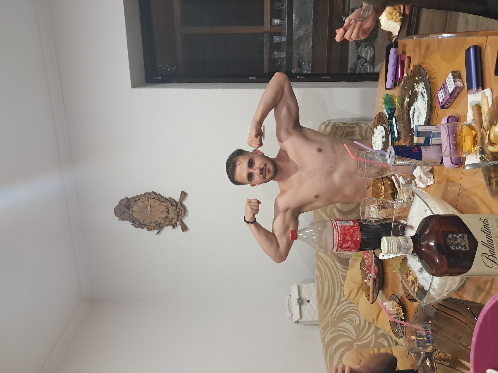 A shirtless man stands flexing both arms. He is indoors, in what appears to be a dining area. A wooden clock is mounted on the wall behind him. He is standing near a table covered with items including bottles (a Coca-Cola bottle and a bottle of Ballantine's scotch whiskey), glasses, plates with food, and disposable lighters and cigarettes. There are people present and eating, at least one of which has a dessert that appears to be cake or ice cream. He has a dark bracelet. The exact location within Gabrovo, Bulgaria, cannot be determined from the image.