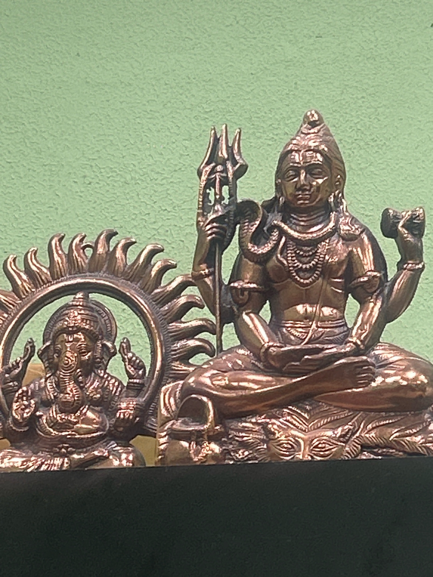The image shows two bronze statues of Hindu deities against a textured light green background. On the left is a statue of Ganesha, recognizable by his elephant head, seated within a radiant halo. To the right is a larger statue of Shiva, depicted with multiple arms, a serpent around his neck, and a trident. Both figures are seated in a meditative pose. The statues rest on a dark base. The scene appears to be indoors, possibly in a home or a religious establishment, but there is no clear indication of its specific location within Almaty, Kazakhstan, or any other city.