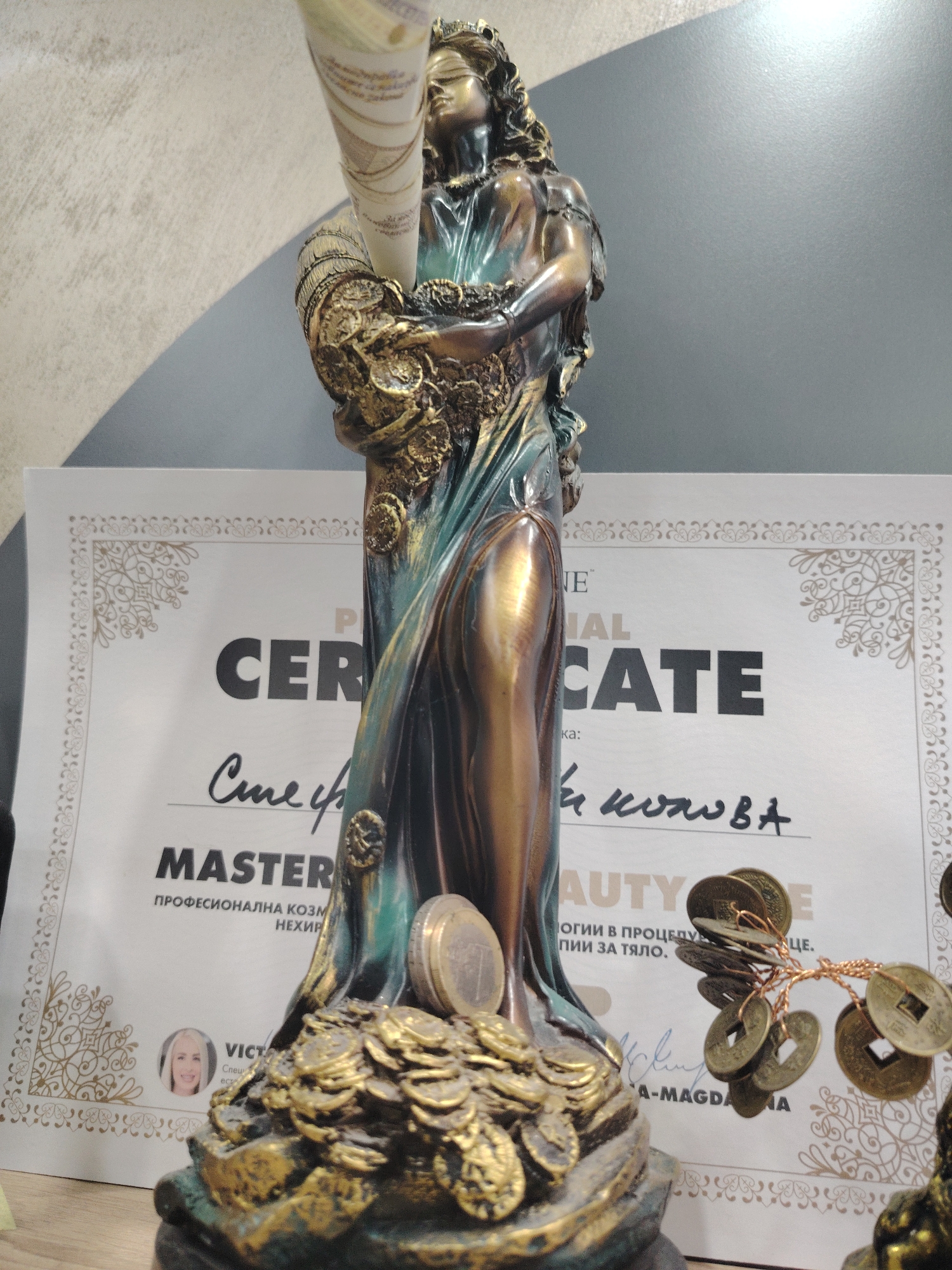 This media file shows a close-up of a decorative statue of Lady Justice, partially obscuring a certificate. The statue is made of bronze and green-patinated metal, depicting a blindfolded woman holding a scroll and a cornucopia overflowing with coins. The base of the statue is also covered in coins, with a euro coin prominently displayed on the statue's drapery. To the right of the statue, a small branch adorned with antique-looking coins is visible.
The certificate behind the statue is a professional certificate, partially obscured by the statue. Visible text on the certificate includes "P", "E", "AL", "CER", "CATE", "MASTER", and "AUTY". Below "MASTER", the text "ПРОФЕСИОНАЛНА КОЗМ НЕХИР" is visible, which translates to "Professional Cosmetology". There is also handwritten text and a signature, and a small portrait of a woman named "VICTA". Another line of text reads "ОГИИ В ПРОЦЕДУР ПИИ ЗА ТЯЛО", possibly referring to procedures for the body. A signature line includes "A-MAGDA".
The setting appears to be indoors, likely a beauty salon or office, given the certificate and the statue's symbolic representation of prosperity. The lighting is artificial and seems to come from the right, casting shadows. The overall mood is one of achievement and perhaps a touch of playful symbolism related to wealth and success in a professional context.