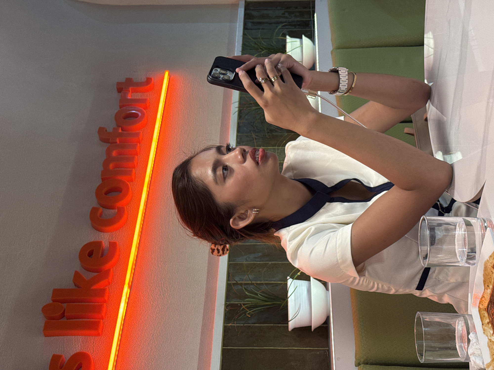 In a softly lit interior, a young woman with dark hair pulled back sits at a white table, engrossed in her smartphone. She wears a white blouse with a navy collar and trim, adorned with gold jewelry on her left hand and wrist. Two glasses of water and a plate of food are visible on the table in front of her. Behind her, a neon sign with orange lettering on a white wall reads "s like comfort." Green padded seating and dark green tiled walls with white planters containing tall, thin plants form the backdrop. The overall ambiance suggests a restaurant or cafe setting, possibly in Davao City, Philippines.