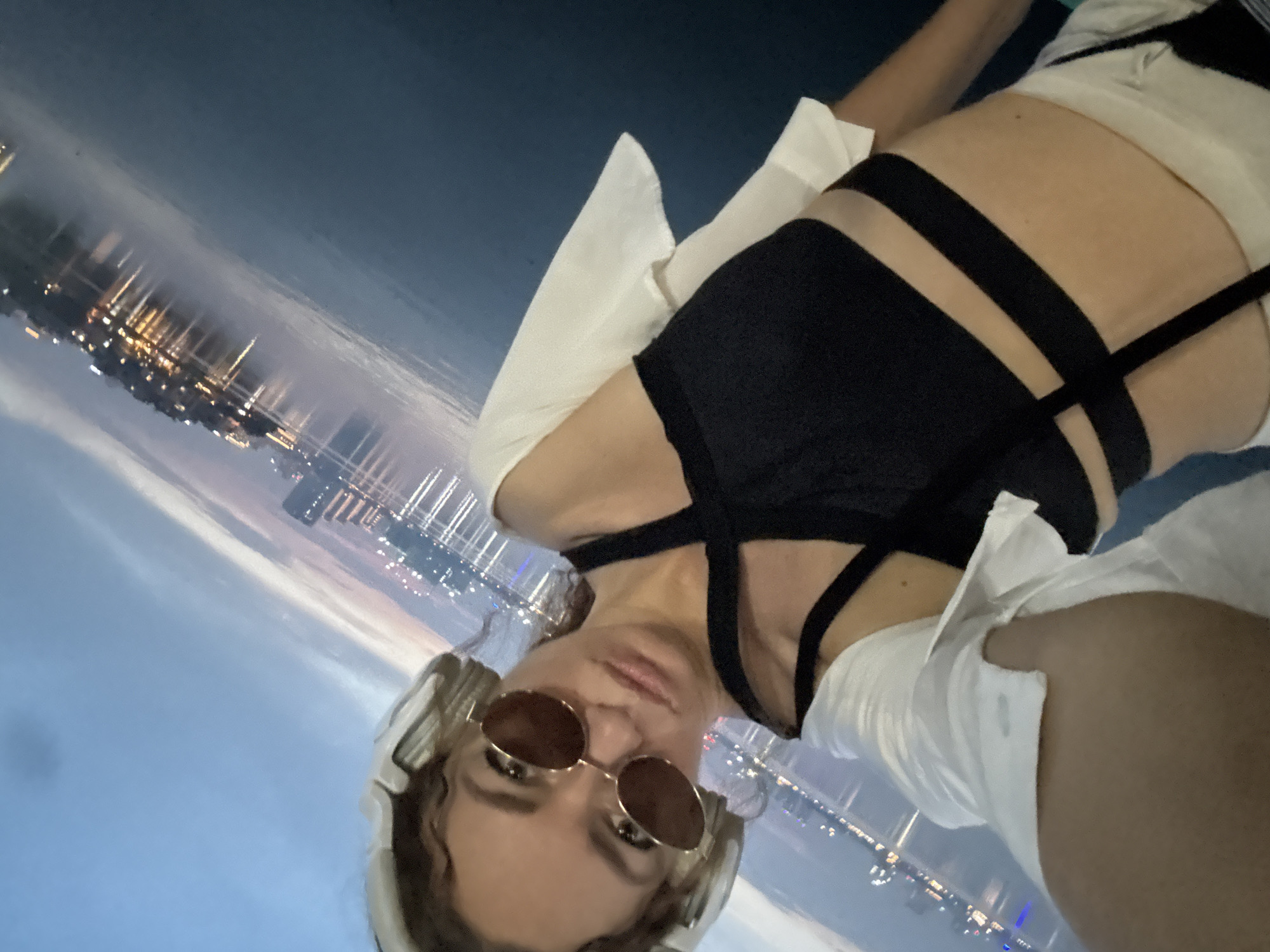 A woman is taking a selfie by the water at dusk in Hanoi, Vietnam. She is wearing a black strappy top, white sleeves, sunglasses, and headphones. The background shows a body of water reflecting the lights of buildings and the sky, typical of Hanoi's lakeside cityscape, such as the area around West Lake or the Red River. The lighting suggests early evening.