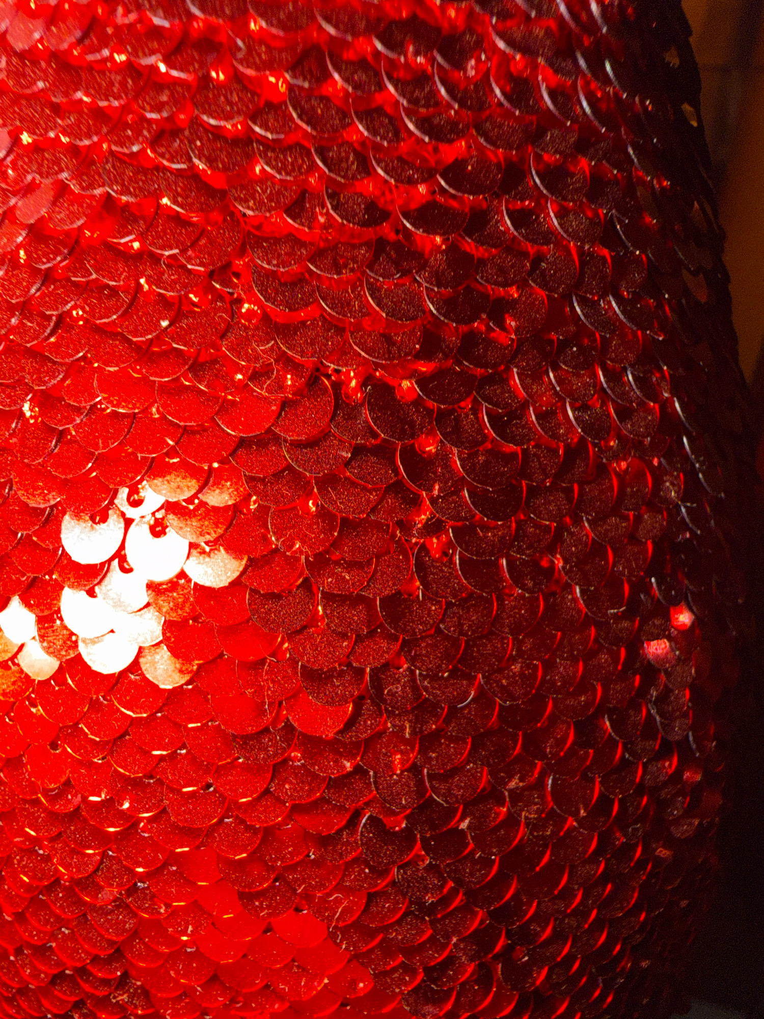 The image is a close-up, high-angle shot of a red sequined surface, likely fabric or an ornament. The sequins are circular and densely packed, overlapping each other in a scale-like pattern. They are predominantly red, with some areas reflecting light brightly, creating highlights and a shimmering effect. The texture is highly reflective and appears to be slightly textured or grainy on some of the individual sequins. The lighting suggests a single light source from the left, casting shadows and defining the contours of the sequins, giving the surface a three-dimensional appearance. The background is dark and out of focus, with some faint reddish-brown tones visible on the right side, suggesting it might be part of the object or its immediate surroundings. There are no people, animals, or discernible landmarks present in the image. The context provided about "Бабініцкі сельскі Савет, Belarus" is not visually represented in the image itself; this detail is external information.