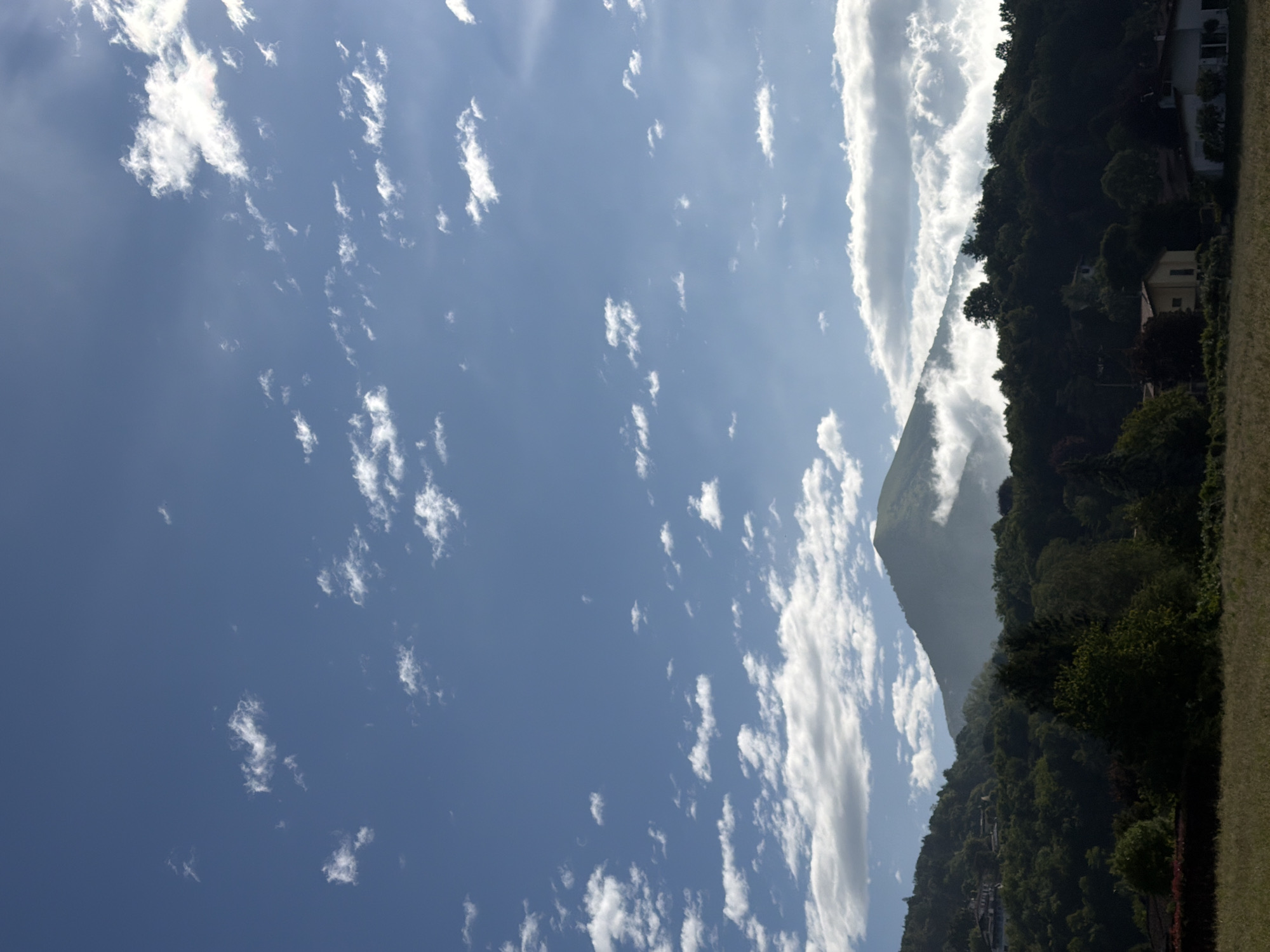 The image shows a view of a mountain, likely near Penzano, Italy, under a partly cloudy sky. The sky is blue with scattered, small, white clouds. A large, vertical cloud formation extends from the ground upwards, obscuring part of the mountain. The mountain is covered in dense vegetation. Some buildings are visible among the trees on the mountain's slope. A grassy area is in the foreground.