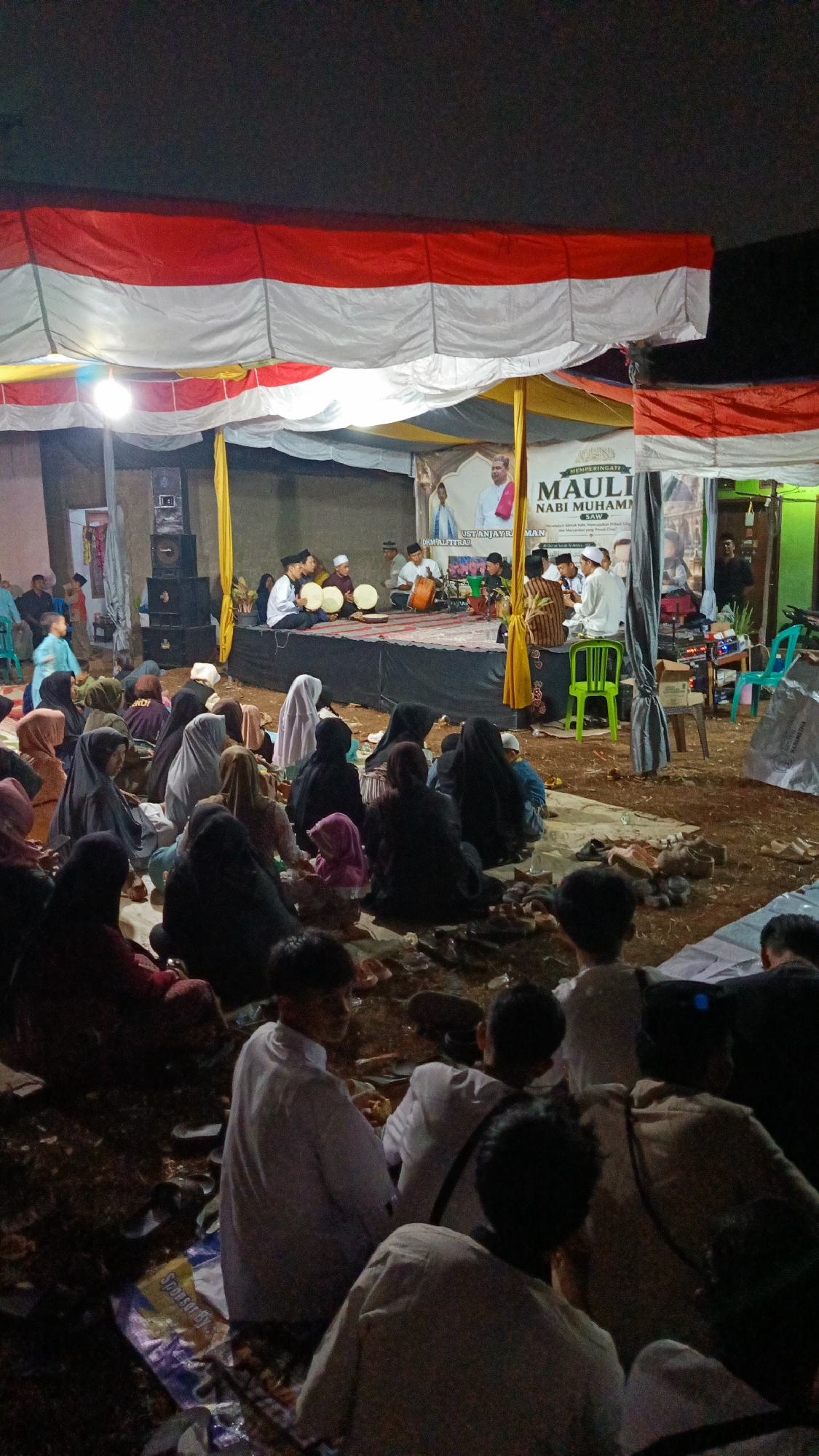 The image depicts an outdoor gathering in Margajaya, Indonesia, likely for a religious event given the banner that reads "MAULIDI NABI MUHAMMAD SAW" which translates to "Birthday of Prophet Muhammad SAW". The scene is set under a canopy draped with red and white Indonesian flags, creating a festive yet solemn atmosphere. A raised platform serves as a stage where several men are seated. Some are playing musical instruments, including what appear to be frame drums, while others are singing or chanting. A banner on the stage features images of religious figures.
In front of the stage, a large audience is seated on the ground, many of whom are women wearing headscarves. The ground is uneven and covered with dirt and scattered items like mats, shoes, and plastic bags. Some individuals are interacting with each other, while others are focused on the performance. The lighting suggests it is evening or nighttime, with artificial lights illuminating the stage and surrounding area. The overall environment appears to be a temporary setup in a residential or open area, with a simple building visible in the background. The presence of speakers and sound equipment indicates a public address system is in use.