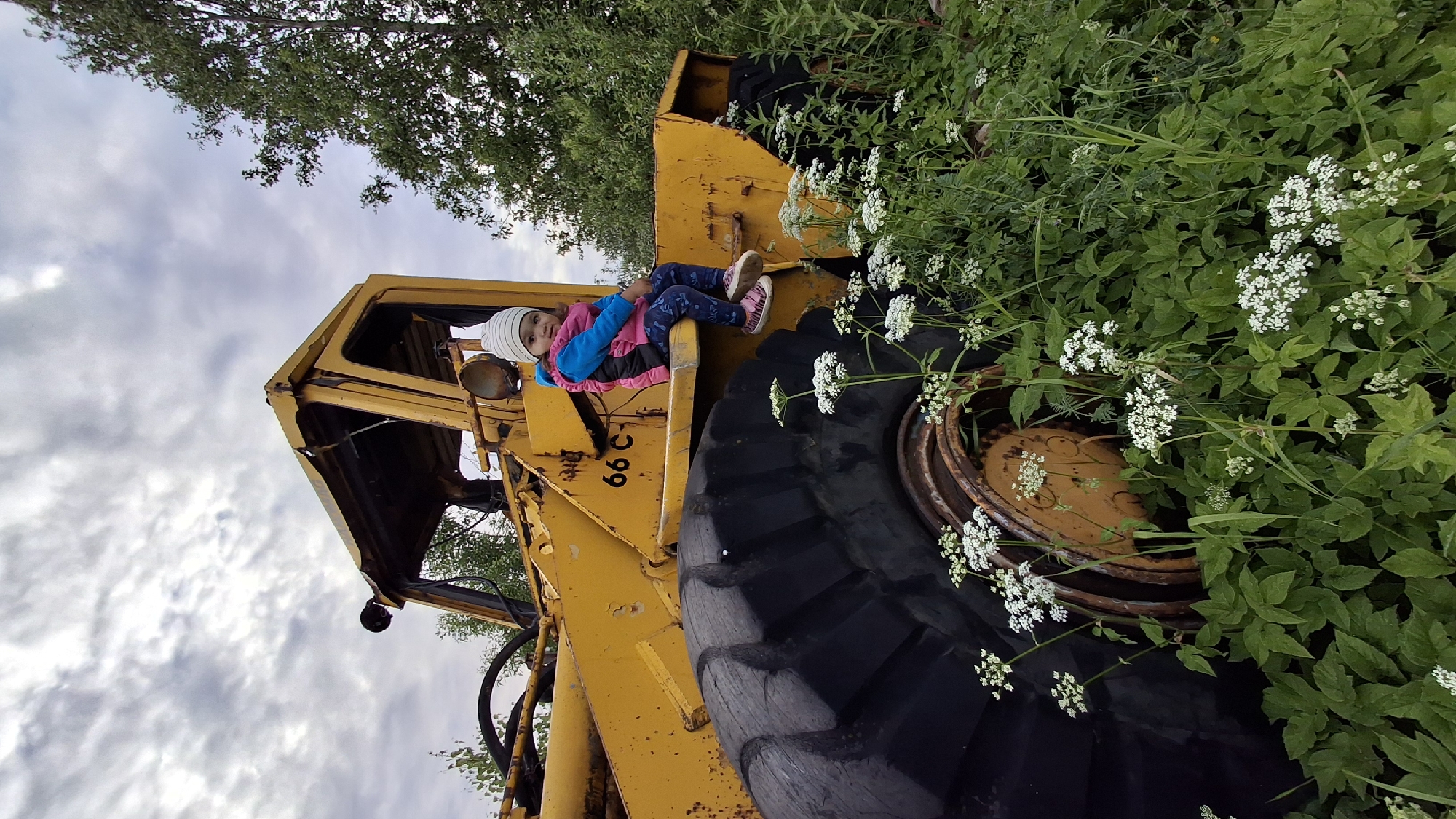 A young child sits on the running board of a yellow, rusted excavator parked amidst overgrown greenery and wildflowers. The child, wearing a striped beanie, a pink vest over a blue shirt, and patterned leggings, looks towards the camera. The excavator's large black tire is visible, as are the words "66 C" marked on its yellow body. The sky above is cloudy. The scene appears to be in a rural or abandoned area, potentially on the outskirts of Innolovo, Russia, given the presence of the excavator and wild vegetation.