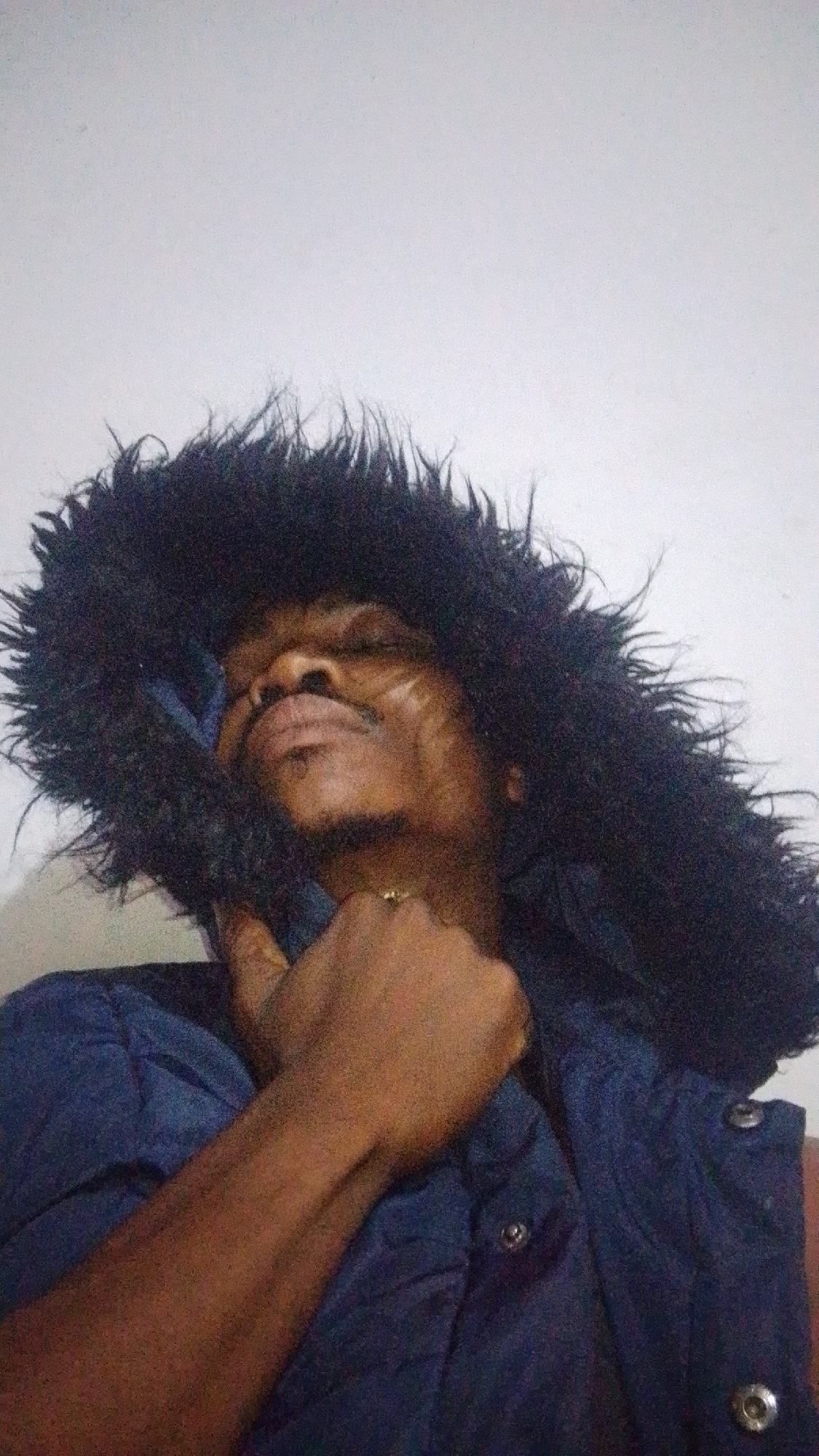 A person is photographed indoors, against a plain white wall. They are wearing a dark blue vest with metallic snap buttons and a large, dark, fluffy hood. The person has dark skin and is looking upwards with their eyes closed, their head tilted back. Their right hand is raised and clasped near their chin, with a ring on their index finger. There are faint marks or lines on their face. The lighting is dim, and the image has a grainy texture.