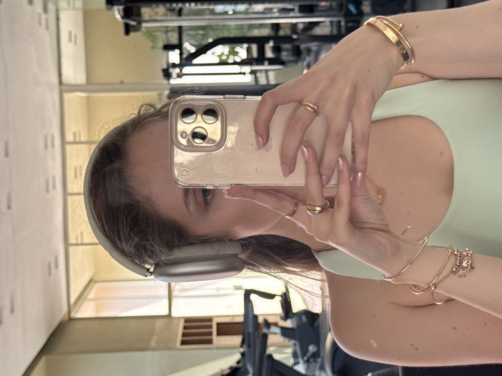 A woman is taking a selfie in what appears to be a gym setting in Mexico City, Mexico. She is holding a smartphone with a clear case, which is obscuring most of her face but revealing one eye and part of her nose. She is wearing headphones over her head. Her left arm is visible with a gold bracelet and what looks like several other rings. Her right arm is also partially visible, wearing two bracelets. Behind her, gym equipment is visible, and a window. The wall behind the woman appears to be a mirror.