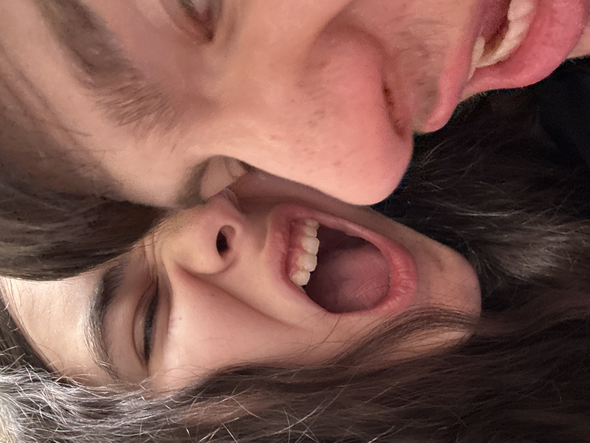 Two individuals are closely positioned, with their faces near each other. One person is positioned upside down with an open mouth and visible teeth. The other person is above them, with their mouth appearing close to the first person's face. Both individuals have light skin tones. The scene is indoors, but without specific elements suggesting the location in Plovdiv, Bulgaria.