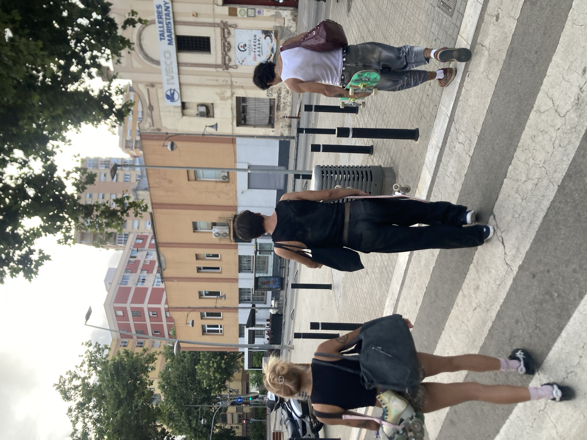 The image shows three individuals walking across a crosswalk in Badalona, Spain. The two individuals in the foreground are walking away from the camera. The person on the left has blonde hair pulled up in a bun and is wearing a black tank top, patterned shorts, pink socks, and black shoes. They are carrying a dark gray tote bag over their left shoulder and a colorful bag in their left hand. The person in the middle is wearing a black tank top and black pants and is carrying a dark bag over their left shoulder. They are also carrying a skateboard in their right hand. The third individual, on the right, is wearing a white tank top, patterned pants, and brown shoes. They are carrying a burgundy shoulder bag over their right shoulder and a green and white skateboard in their right hand.
The background features a street scene with buildings, trees, and streetlights. One building has prominent signage that reads "IVECO TALLERES MARISTANY," indicating a location in Badalona. The weather appears overcast. The crosswalk is marked with white and gray stripes. A motorcycle is parked on the left side of the image.