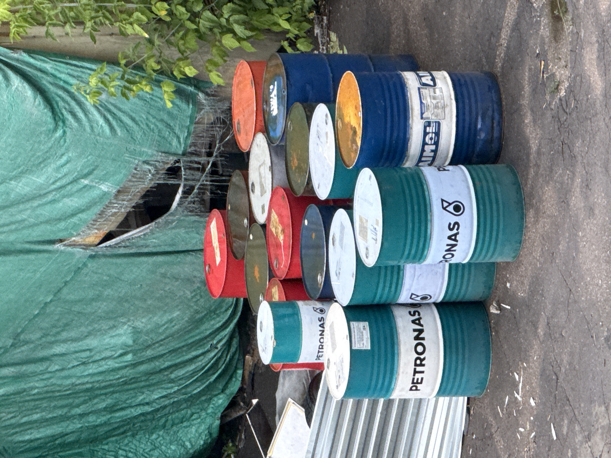 A collection of oil drums are stacked and arranged on a concrete surface. Some of the drums are painted red, orange, green, and blue. Several drums bear the "PETRONAS" logo, indicating they likely contain petroleum products. A large, ripped green tarp hangs in the background, partially obscuring a structure. Lush green foliage grows on the right side of the image, adjacent to a building facade. The scene appears to be outdoors, possibly in a storage or industrial area. There are no people or animals visible in the image. The ground is paved with asphalt and shows some scattered debris. The image does not provide any specific information to suggest a location in Moscow, Russia.