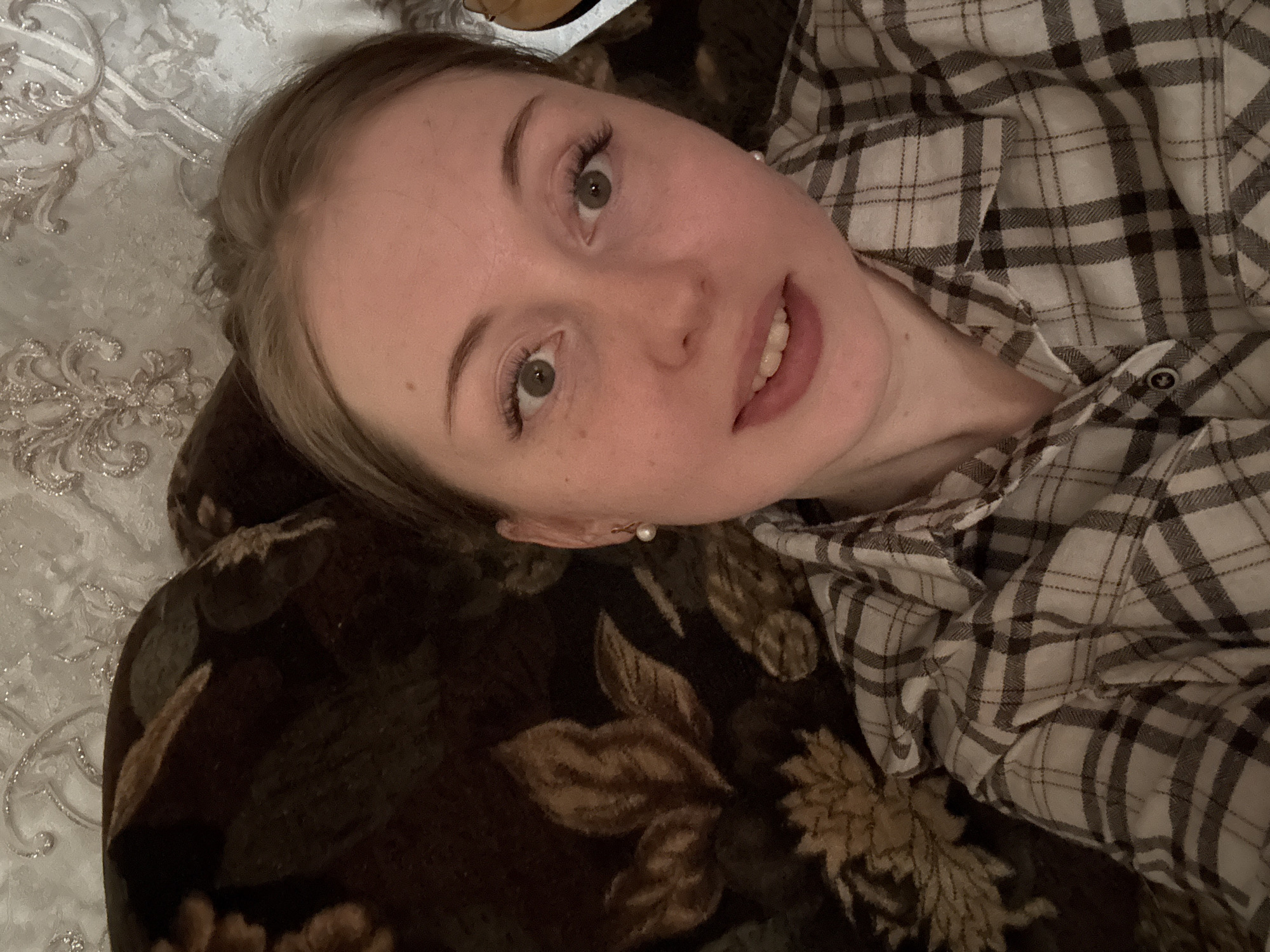 A close-up, slightly angled photograph depicts a Caucasian woman lying down. She has light skin, blonde hair pulled back, and green eyes. She is wearing a grey and black plaid shirt. A pearl earring is visible on her left ear. The woman is lying on a floral print brown couch. Behind the couch is a patterned cream-colored bedspread. The location and specific actions are unknown.