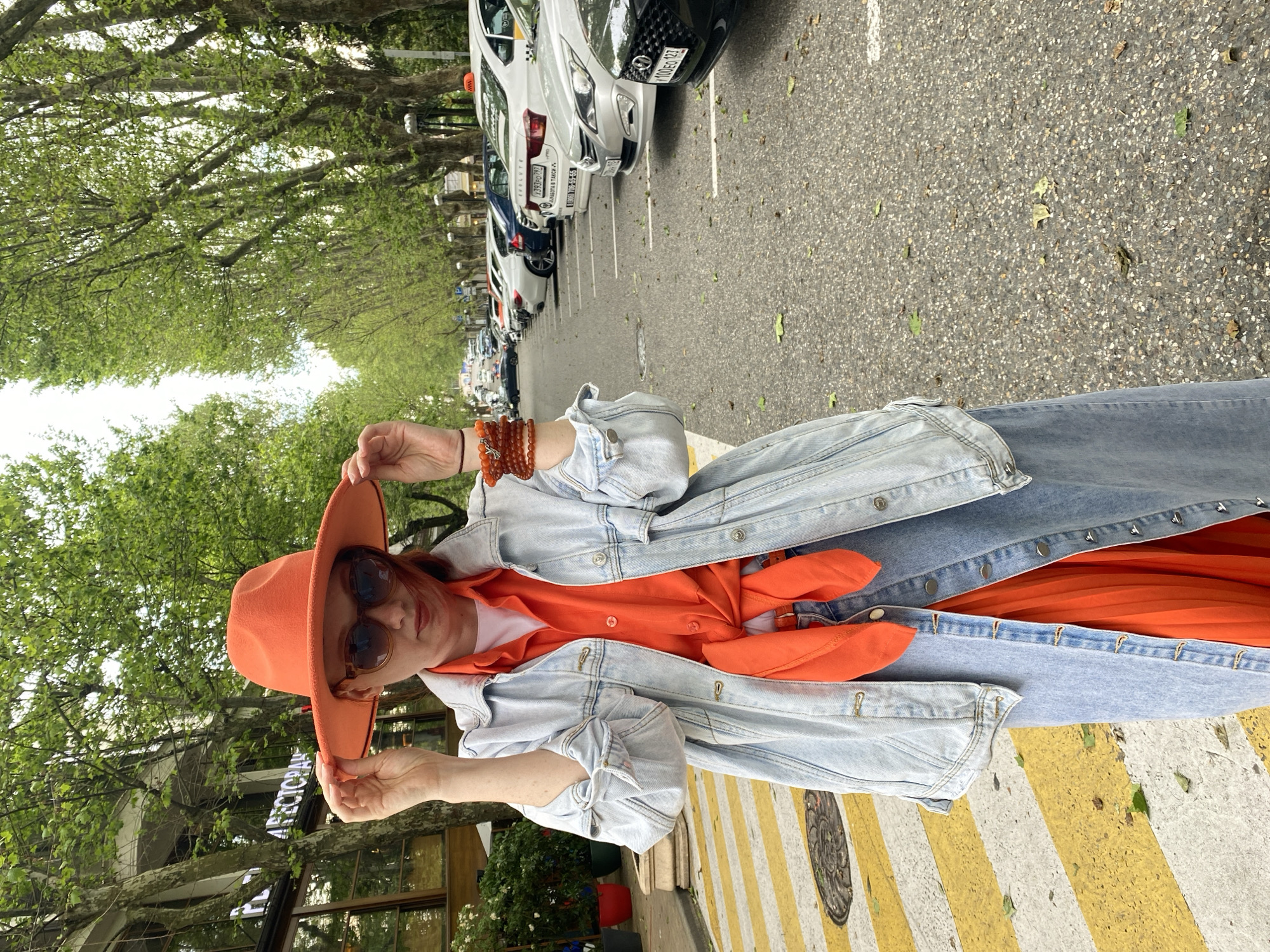 A woman with reddish hair, sunglasses, and an orange hat stands on a street in Sochi, Russia. She is wearing a denim jacket over an orange shirt or dress. Cars are parked along the side of the road, and trees line the street. Yellow and white stripes are painted on the road.
