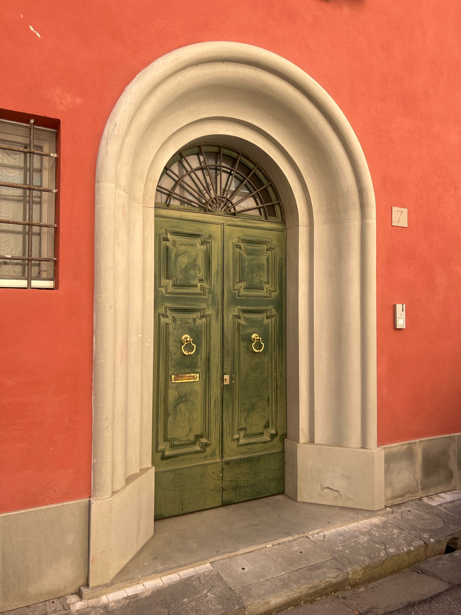 The image shows a green double door with ornate carvings and brass door knockers, set within an arched stone entryway. The wall surrounding the door is painted a vibrant coral red. To the left of the door is a window with a metal grate. A house number "7" is visible on the wall to the right of the entryway, along with a small white intercom unit below it. The entryway and door are situated on a stone sidewalk with a curb leading to a street. The scene depicts a section of a building facade in a European city, possibly Pisa, Italy, given the architectural style.