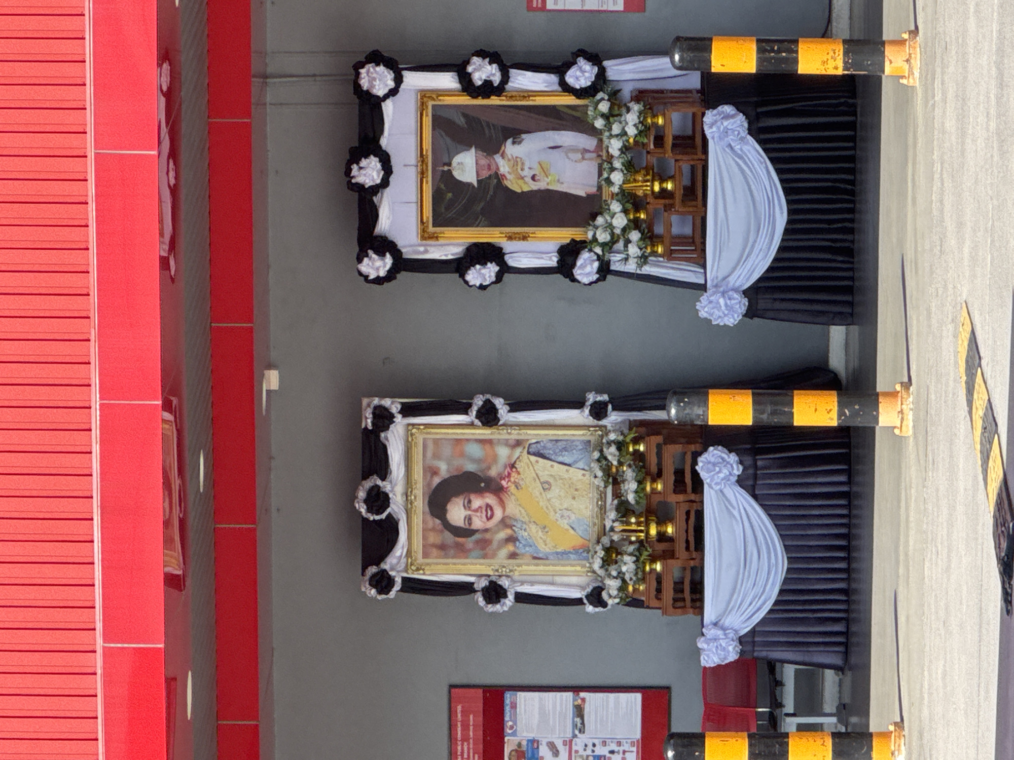 This image shows two framed portraits displayed on tiered wooden stands in front of a building with a red corrugated metal facade. The portrait on the left depicts a woman wearing a traditional Thai outfit with a yellow sash. The portrait on the right shows a person in a white military uniform with a helmet. Both portraits are adorned with black and white fabric drapery and artificial white flowers. In front of each portrait, there are arrangements of white flowers and gold pedestals. The overall setting appears to be an outdoor area, possibly in a public space, with black and yellow bollards visible in the foreground. The context of the display suggests a memorial or a somber occasion.