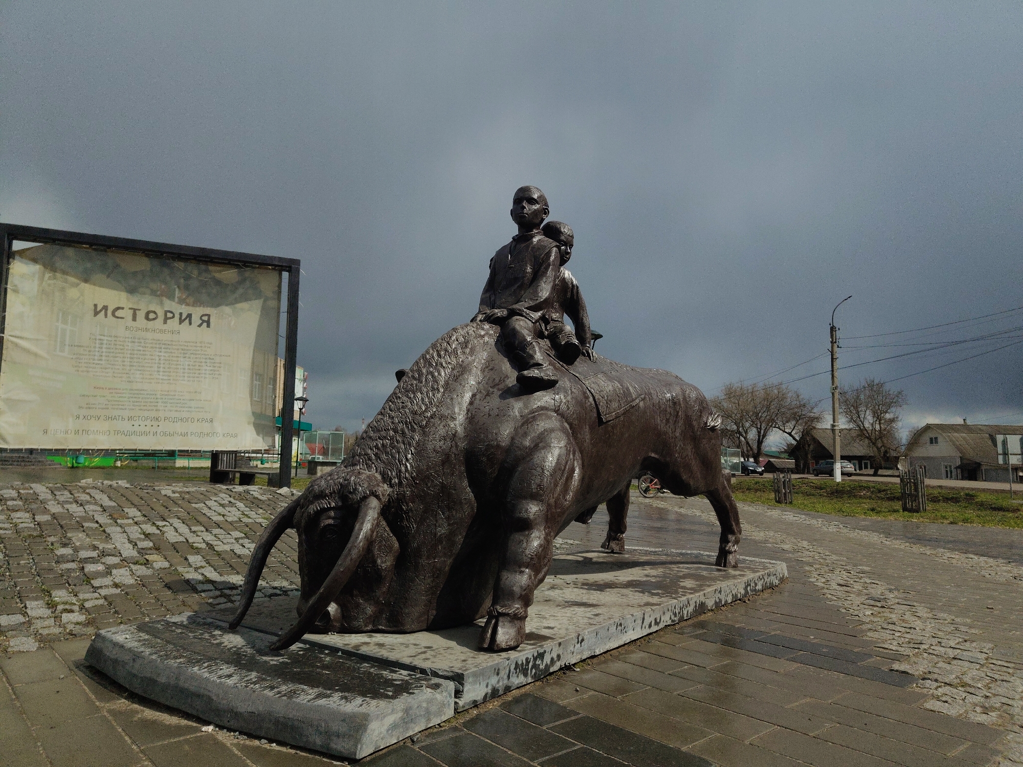 A bronze-colored statue dominates the foreground, depicting a large bull or bison with two children riding on its back. The animal is rendered in a powerful, grounded stance, with its head lowered and prominent horns curving forward. The first child, seemingly a boy, sits upright in front, appearing to hold reins, while a second, smaller child sits directly behind him. The statue rests on a rectangular concrete base, which shows signs of dampness.
To the left of the statue stands a large outdoor information board or sign, framed in dark metal. The text on the sign is in Cyrillic, reading "ИСТОРИЯ" (HISTORY) and "ВОЗНИКНОВЕНИЯ" (ORIGIN/EMERGENCE) at the top. Further down, it states: "Я ХОЧУ ЗНАТЬ ИСТОРИЮ РОДНОГО КРАЯ" (I WANT TO KNOW THE HISTORY OF MY NATIVE LAND) and "Я ЦЕНЮ И ПОМНЮ ТРАДИЦИИ И ОБЫЧАИ РОДНОГО КРАЯ" (I VALUE AND REMEMBER THE TRADITIONS AND CUSTOMS OF MY NATIVE LAND). The board's surface appears slightly weathered or translucent, revealing faint outlines of structures behind it.
The setting is an outdoor paved area, with cobblestone and concrete slabs visible on the ground, which appears wet or damp. The sky is overcast and grey, suggesting a cloudy day, possibly after rain. In the background, leafless trees, residential-style buildings, a street light, and power lines are visible, indicating a town or village environment. A red bicycle wheel can be faintly seen near the hindquarters of the statue, belonging to a parked bicycle. The overall scene is calm and somewhat somber due to the weather and dark tones of the statue.