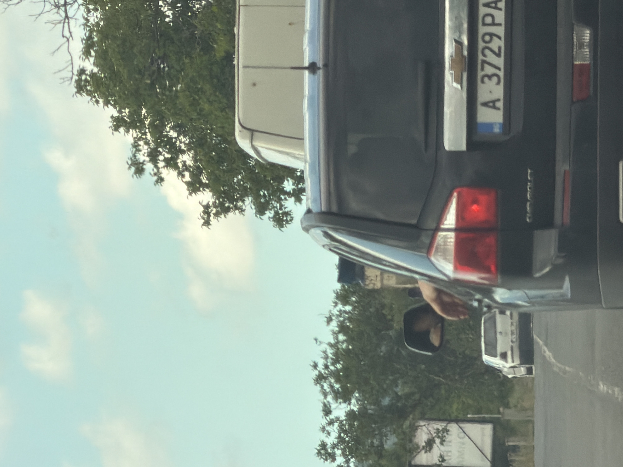 The image captures a scene from within a vehicle traveling on a road in Aytos, Bulgaria, as indicated by the license plate "A 3729 PA" on the vehicle in the foreground. The vehicle in the foreground, a dark SUV, has its rear license plate prominently displayed. To the left, a white sedan is visible further down the road. The environment appears to be a suburban or rural area with trees and vegetation lining the roadside. The sky is a light blue with scattered clouds. A person's arm is visible resting on the driver's side window of the foreground vehicle, suggesting the photo was taken from inside the moving vehicle. The overall lighting suggests it is daytime.