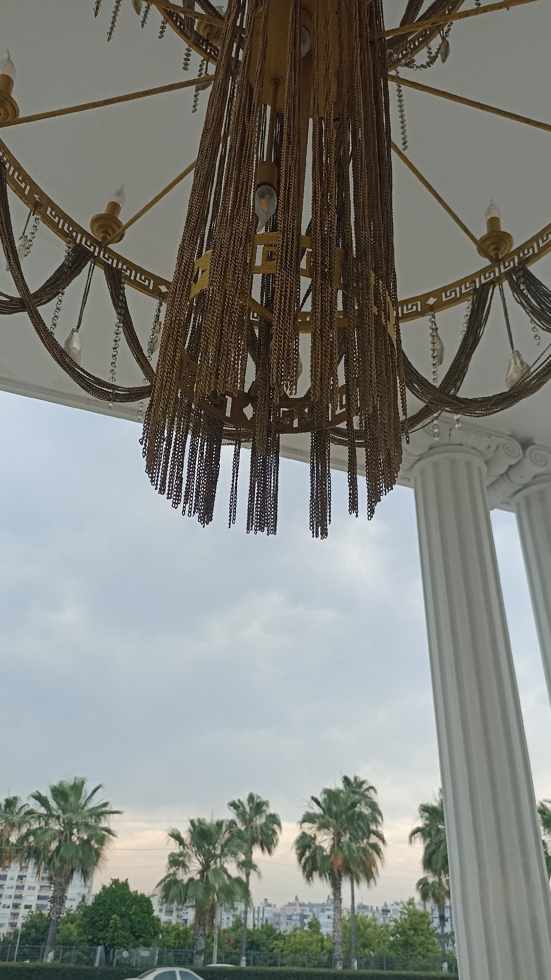 The image depicts an ornate, gold-colored chandelier hanging from a white ceiling. The chandelier is adorned with numerous thin gold chains that drape downwards, creating a textured effect. Several small, clear bulbs are visible within the chandelier's structure. The ceiling appears to be part of an architectural structure, possibly a covered walkway or portico, as suggested by the presence of large, white classical columns to the right of the frame.
In the background, the scene transitions to an outdoor environment under a cloudy sky. A line of palm trees is visible, interspersed with various buildings and structures, suggesting an urban or suburban setting. The overall impression is one of a well-appointed outdoor or semi-outdoor area, likely in a warm climate. The location is identified as Yenişehir, Turkey. No people are visible in the image.