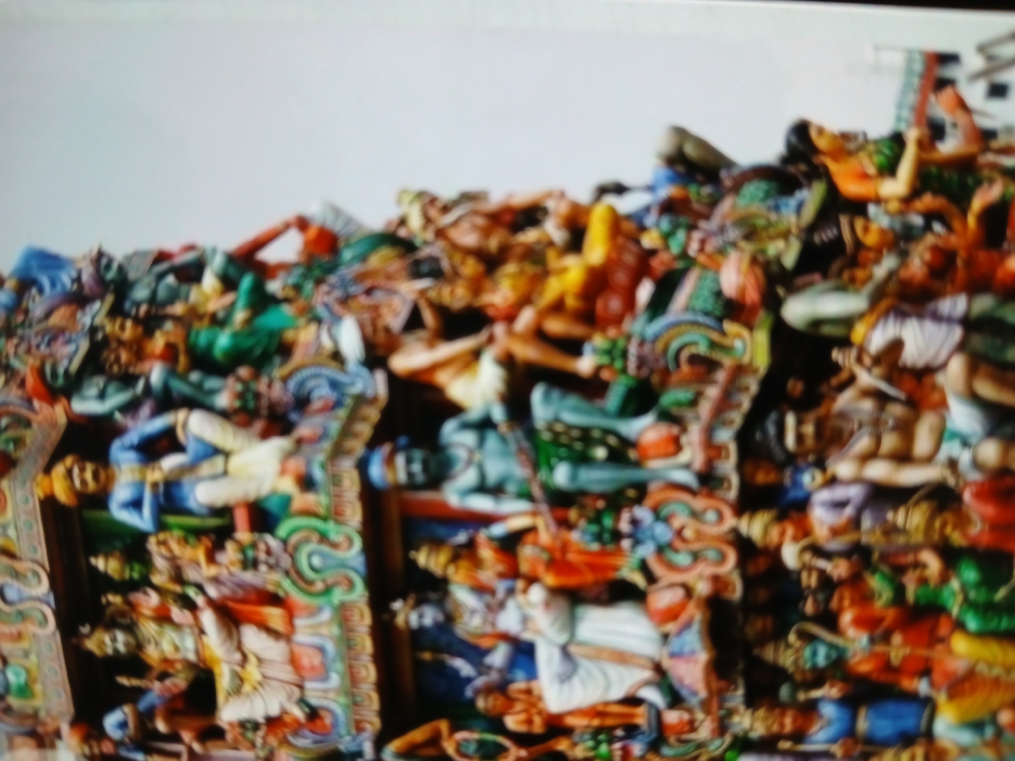 The image depicts a close-up view of intricately carved and brightly painted sculptures, likely from a Hindu temple. The figures are predominantly human and possibly divine, adorned with ornate garments and headdresses. The scene suggests a densely populated architectural element, potentially a gopuram or a ceiling panel in a temple. Given the prompt, the likely location is Bengaluru, India, suggesting this is a detail from a temple in that city.