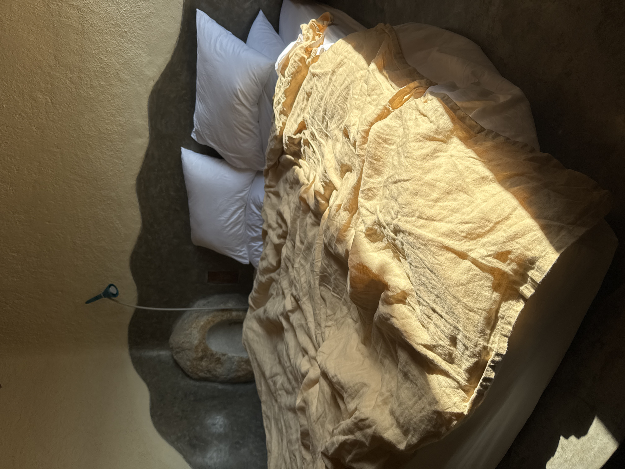 An overhead shot captures a sunlit bedroom in Bac Nha Trang Ward, Vietnam, highlighting a bed adorned with white pillows and a rumpled, light beige or golden-yellow duvet. Intense natural light streams into the room, creating a dramatic interplay of bright illumination and deep, organic shadows. The duvet's textured, linen-like fabric is emphasized by the light, showing numerous wrinkles and folds.
Three plump white pillows are arranged at the head of the bed, resting against a textured, earthy-toned wall. The wall exhibits a rough, stucco-like finish, with its golden-brown hue varying from light in sunlit areas to darker tones in shadow.
On the dark, textured floor beside the bed, a round, rough-hewn stone basin or bowl is visible, partially obscured by shadows. A thin wire extends from the basin area towards the wall, connecting to a small, light blue-green object, possibly an electrical fixture or sensor.
Wavy, irregular shadows are cast across the floor and the lower part of the wall, suggesting sunlight filtering through an unusual opening or external elements. The sharp, directional quality of the light indicates either early morning or late afternoon. The scene evokes a serene and minimalist aesthetic, focusing on natural textures, warm earth tones, and the dynamic effects of sunlight within this rustic interior. No people or visible text are present.