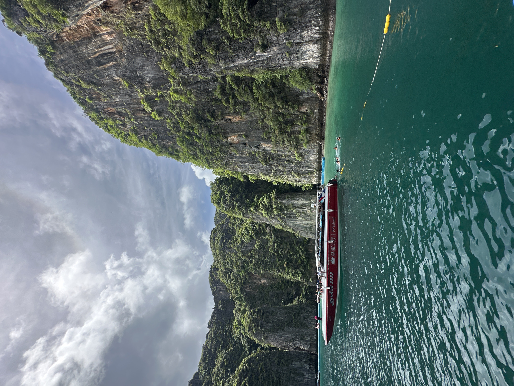 A red and white boat with the text "PP Island" and "3333" is docked next to a steep, lush green cliff face. Several people are visible on the boat, and a few are swimming in the turquoise water. A white rope with yellow buoys stretches across the water, possibly marking a swimming area or boundary. The sky is partly cloudy, with some bright patches and some darker, more dramatic clouds. The sun appears to be casting dappled light on the water, suggesting it is midday. The scene is likely a tourist destination, given the presence of a tour boat and swimmers. The overall mood is serene and adventurous, with the majestic natural scenery contrasting with the recreational activities. The location appears to be tropical, given the vegetation and the clear, warm-looking water.