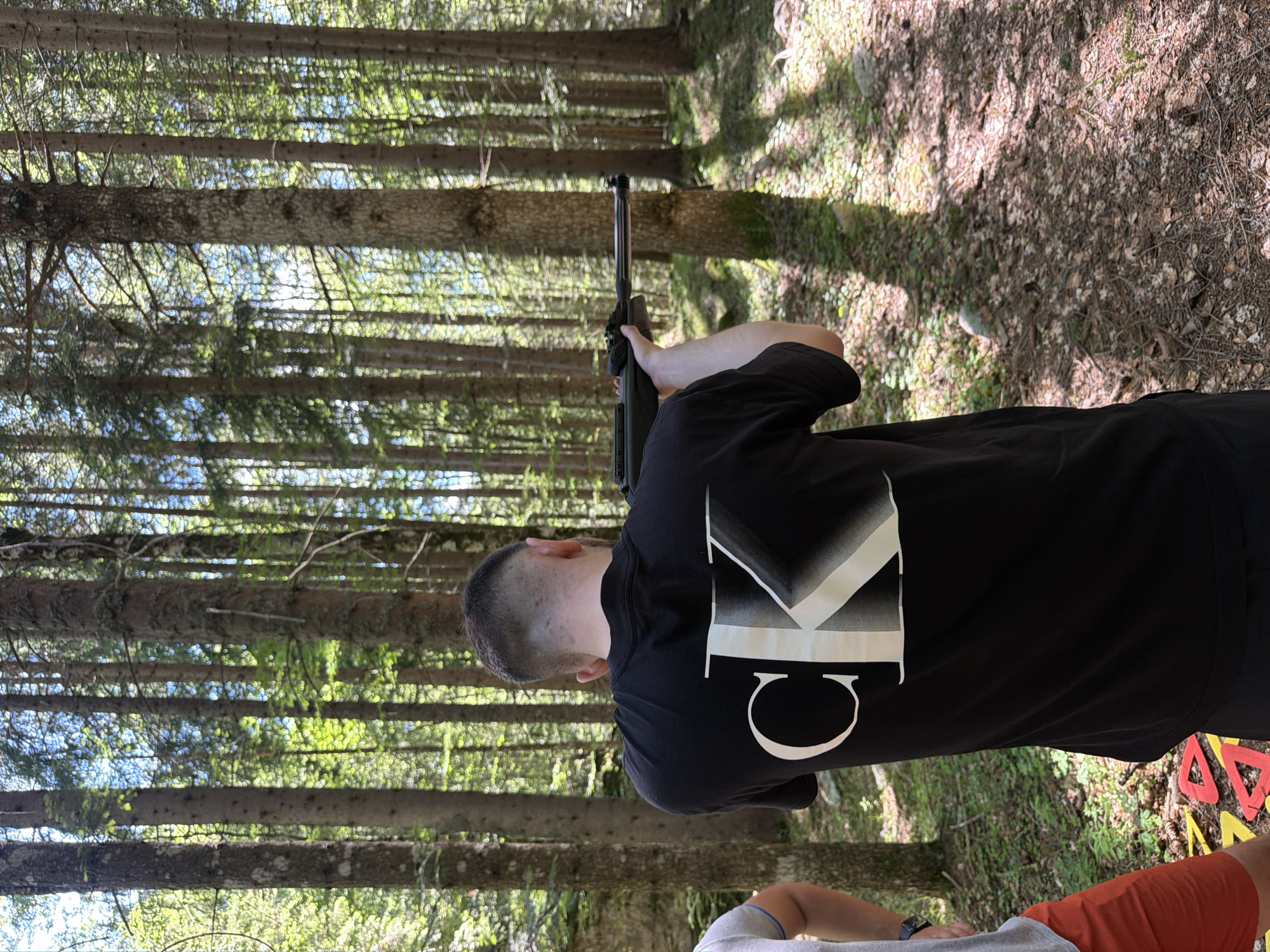 In Govedartsi, Bulgaria, a person in a black t-shirt with a white "CK" logo is aiming a dark-colored rifle through a dense forest. The person's back is to the camera, and their head is turned slightly to the right. The forest floor is covered in pine needles and fallen leaves, with a scattering of small rocks. Sunlight filters through the tall pine trees, creating dappled shadows on the ground. Another person, partially visible on the left side of the frame, is wearing a grey t-shirt and orange shorts. Colorful triangular targets are laid out on the ground near their feet. The scene appears to be an outdoor recreational activity.