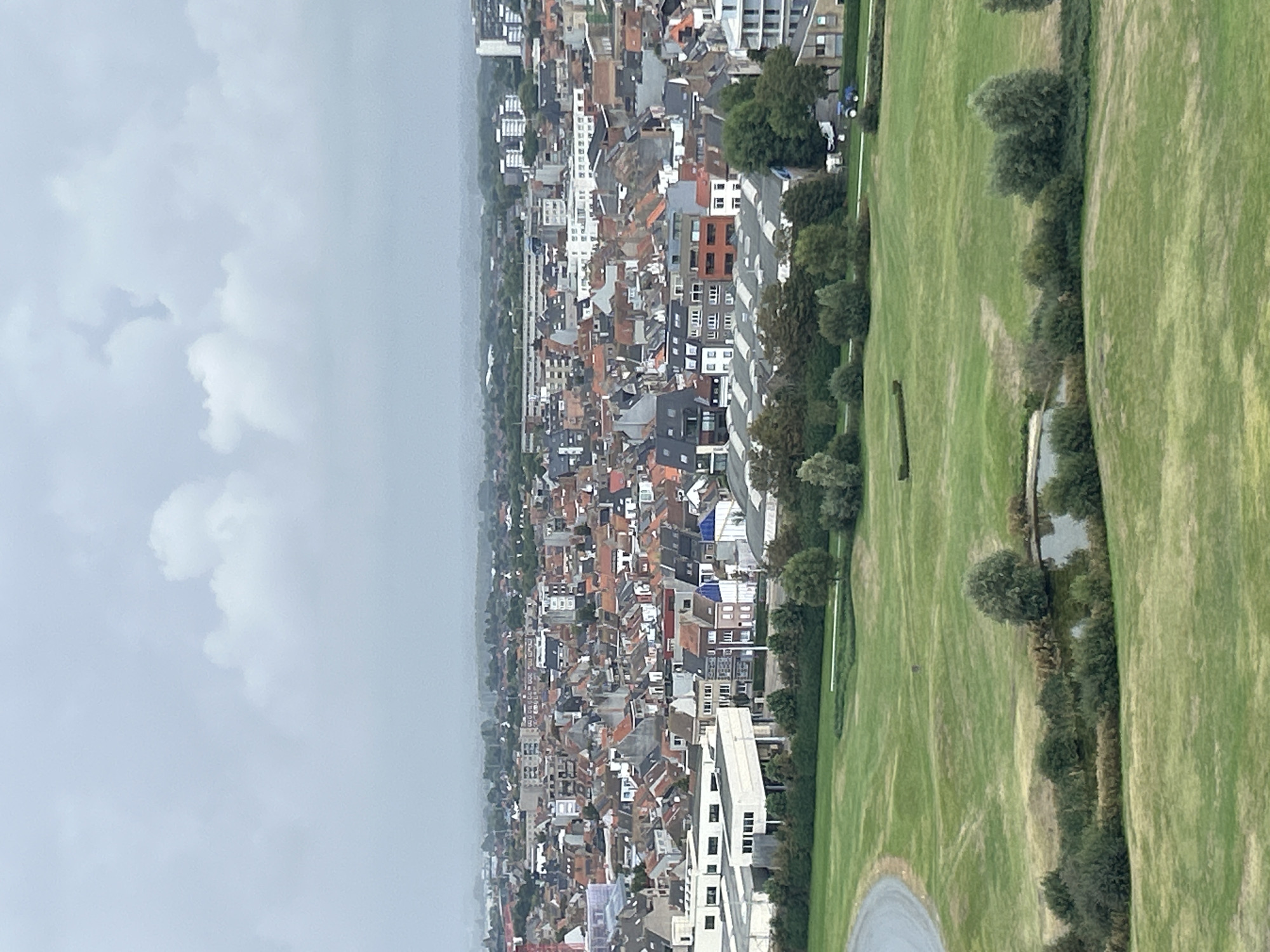 The image depicts a panoramic view of the city of Ostend, Belgium, under an overcast sky. In the foreground, a lush green lawn with rolling hills and a small pond is visible. A paved pathway curves into the frame from the left.
Beyond the park, a dense urban landscape unfolds, characterized by a multitude of buildings with red-tiled roofs. The architecture suggests a European city with varying building heights and styles, ranging from traditional houses to more modern structures. In the mid-ground, a prominent white building complex stands out, featuring a multi-story design with numerous windows. Further in the distance, taller buildings and a more scattered urban fabric extend towards the horizon, which is obscured by mist or haze.
A small blue tractor is visible on a path in the middle ground, suggesting some activity or maintenance in the area. The overall scene conveys a sense of urban life integrated with green spaces, observed from an elevated vantage point. The time of day appears to be daytime, given the ambient light, but the cloudy conditions subdue the intensity of the light.