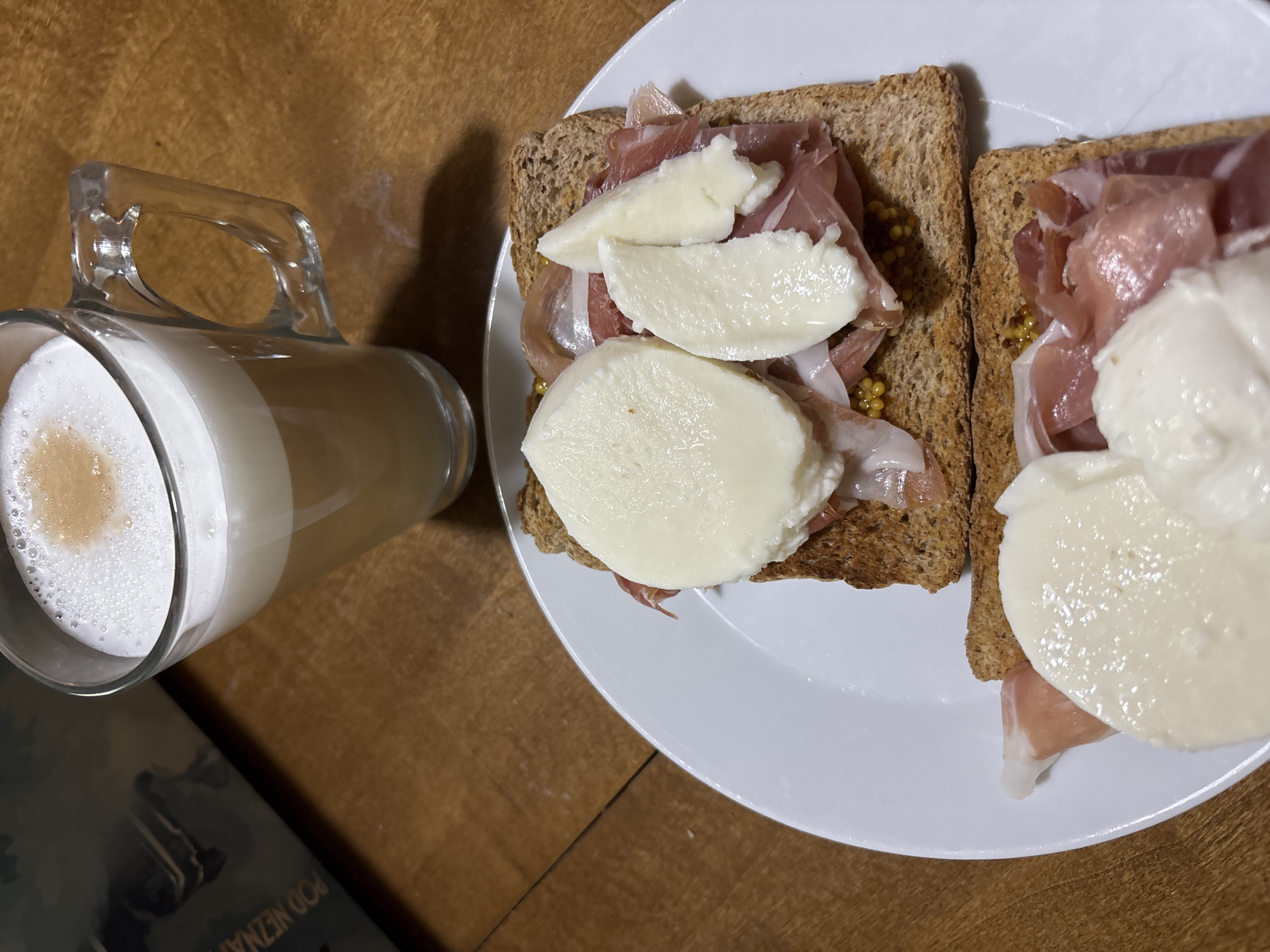 This media file depicts a close-up, overhead view of two open-faced sandwiches with prosciutto and mozzarella, served on whole wheat toast, resting on a white plate. The sandwiches are topped with slices of fresh mozzarella and appear to have a spread of mustard beneath the prosciutto. Next to the plate is a clear glass mug filled with a layered coffee drink, likely a latte or cappuccino, with a frothy top. A book or magazine with text visible lies on the wooden table in the foreground, partially obscuring the mug. The overall scene suggests a light meal or snack. The setting is a wooden surface, consistent with a table. The lighting suggests it might be daytime, though no specific time can be definitively determined. No people are visible in the image. Visible text on the book includes "POD NEZNÁMÝ..." which is likely part of a title or phrase. The location context of Červená Píska, Czechia is provided but not visually evident within the image itself.