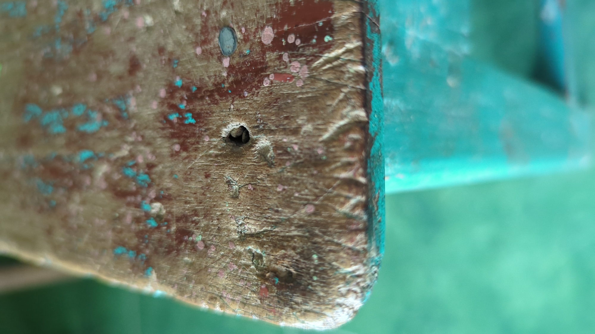 The image shows a close-up, low-angle shot of a weathered, wooden surface with chipped paint. The wood grain is visible, and there are remnants of red and teal paint. A small, dark hole is present in the wood, and a metallic disc or nail head is also visible to the right of the hole. The surface appears to be the edge of a table or desk, with a teal-colored surface below. The background is a blurry, greenish-blue, suggesting an outdoor or lightly colored indoor environment. There are no people or animals in the image. The scene is located in Penjor, Indonesia, and is a forensic depiction of an object's surface.