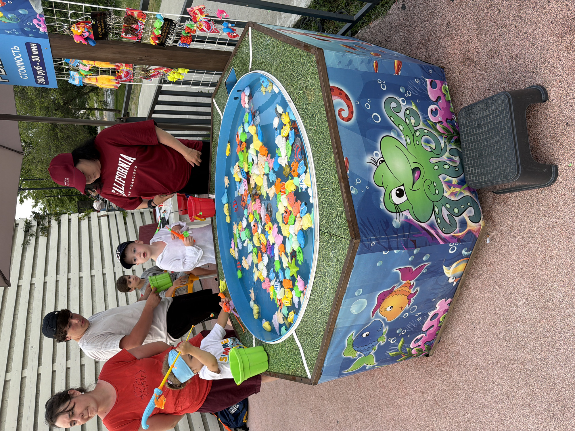 In Sochi, Russia, a children's fishing game is set up outdoors. A woman in a red shirt stands to the left, her gaze directed downwards. Beside her, a teenage boy in a grey t-shirt and black shorts holds a green bucket, looking at a young boy in a white shirt and blue headband who is actively playing the game with a fishing rod. Another young boy, wearing a white long-sleeved shirt and a dark baseball cap, also participates.
The game features a hexagonal pool filled with water, teeming with colorful plastic fish and sea creatures. The sides of the game are decorated with vibrant underwater scenes, including cartoonish fish and a large green octopus. A dark plastic stool sits in front of the game for participants. In the background, a display rack holds various keychains, suggesting they are prizes for the game. The ground is covered in a pinkish textured material, and a white wooden wall forms the backdrop. A black umbrella provides shade over the area. The overall scene captures a lively moment of outdoor recreation.