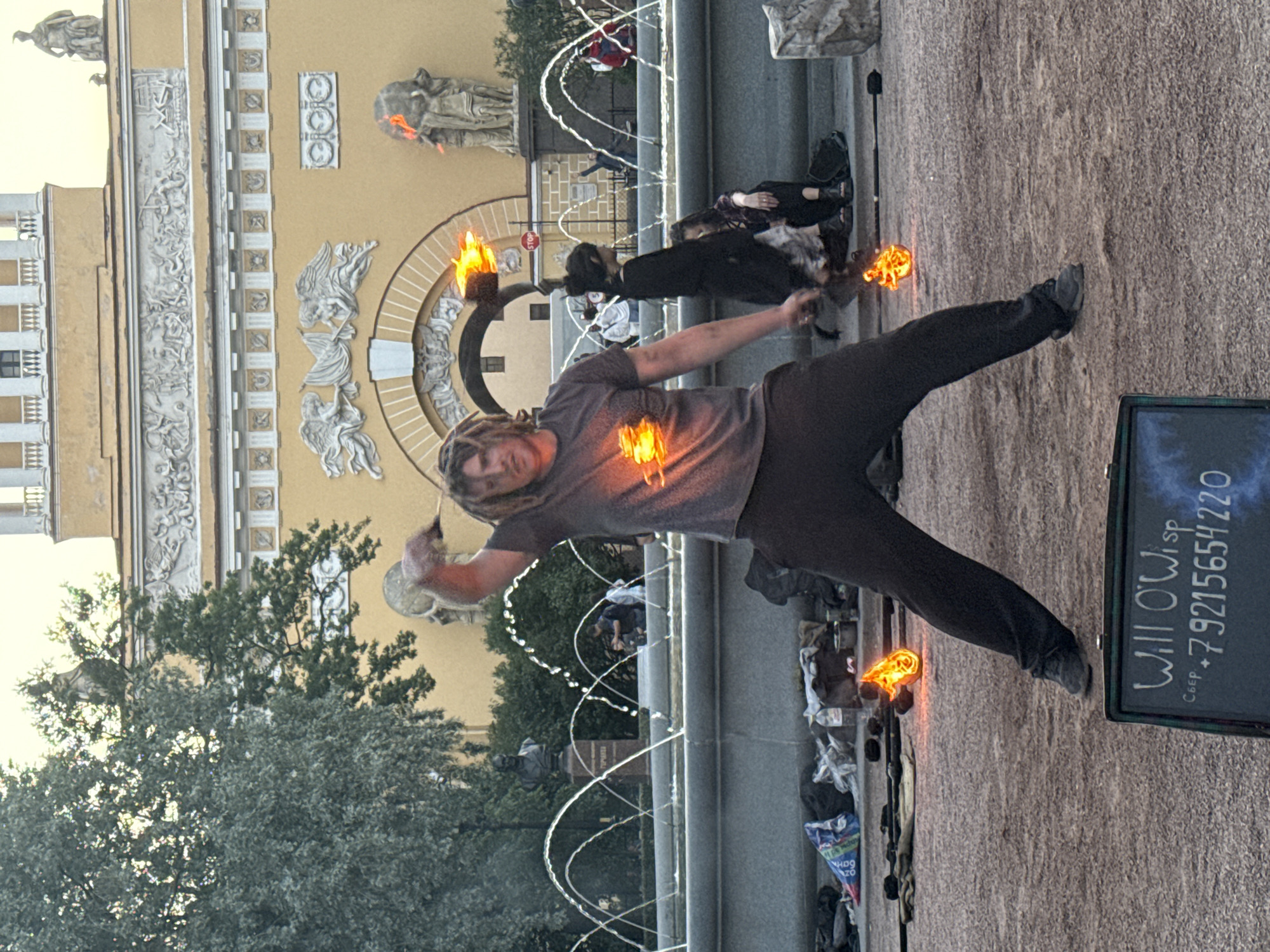 This image depicts a street performer in Saint Petersburg, Russia, performing with fire in front of the main building of the General Staff Building on Palace Square. The performer, a man with dreadlocks wearing a grey t-shirt and dark pants, is in the foreground, holding a flaming object in his right hand and appearing to spin it. Flames are also visible on his chest. In the background, a large fountain sprays water, and several people are gathered, some watching the performance. The architecture of the General Staff Building, with its elaborate classical decorations, is visible behind the fountain. The ground is a sandy, gravelly surface. A black sign with white lettering, displaying "Will O'Wisp" and a phone number, is placed in the lower right corner of the frame.