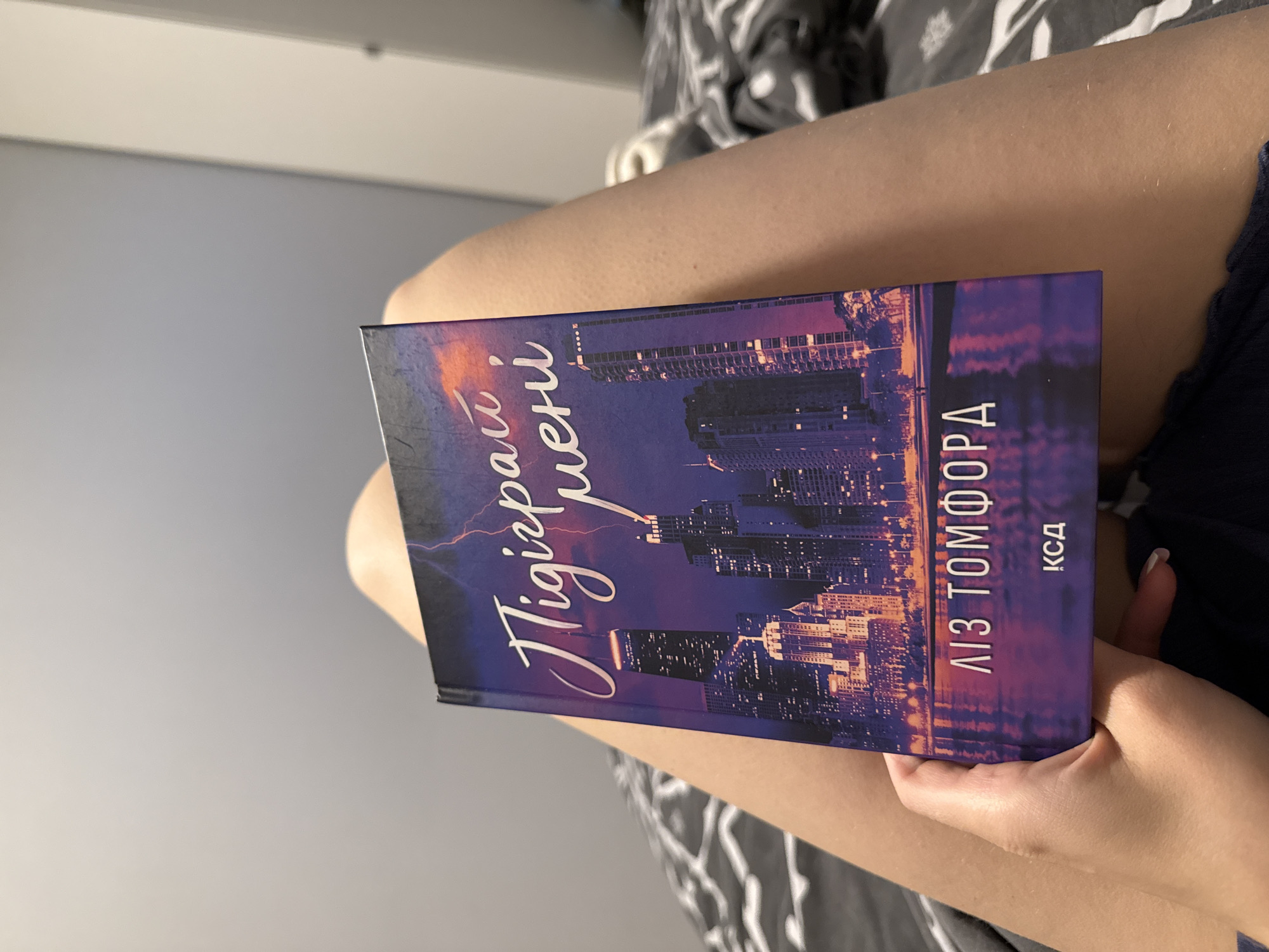 A book with a purple cover featuring a nighttime cityscape with lightning strikes is being held by a person whose legs are visible. The book's title, written in a script font, is "Підіграй мені". Below the title, the author's name "ЛІЗ ТОМФОРД" and a logo "КСП" are present. The person's hand is holding the bottom right corner of the book. The background consists of bedding. The image does not contain any information about the city of Łódź, Poland.