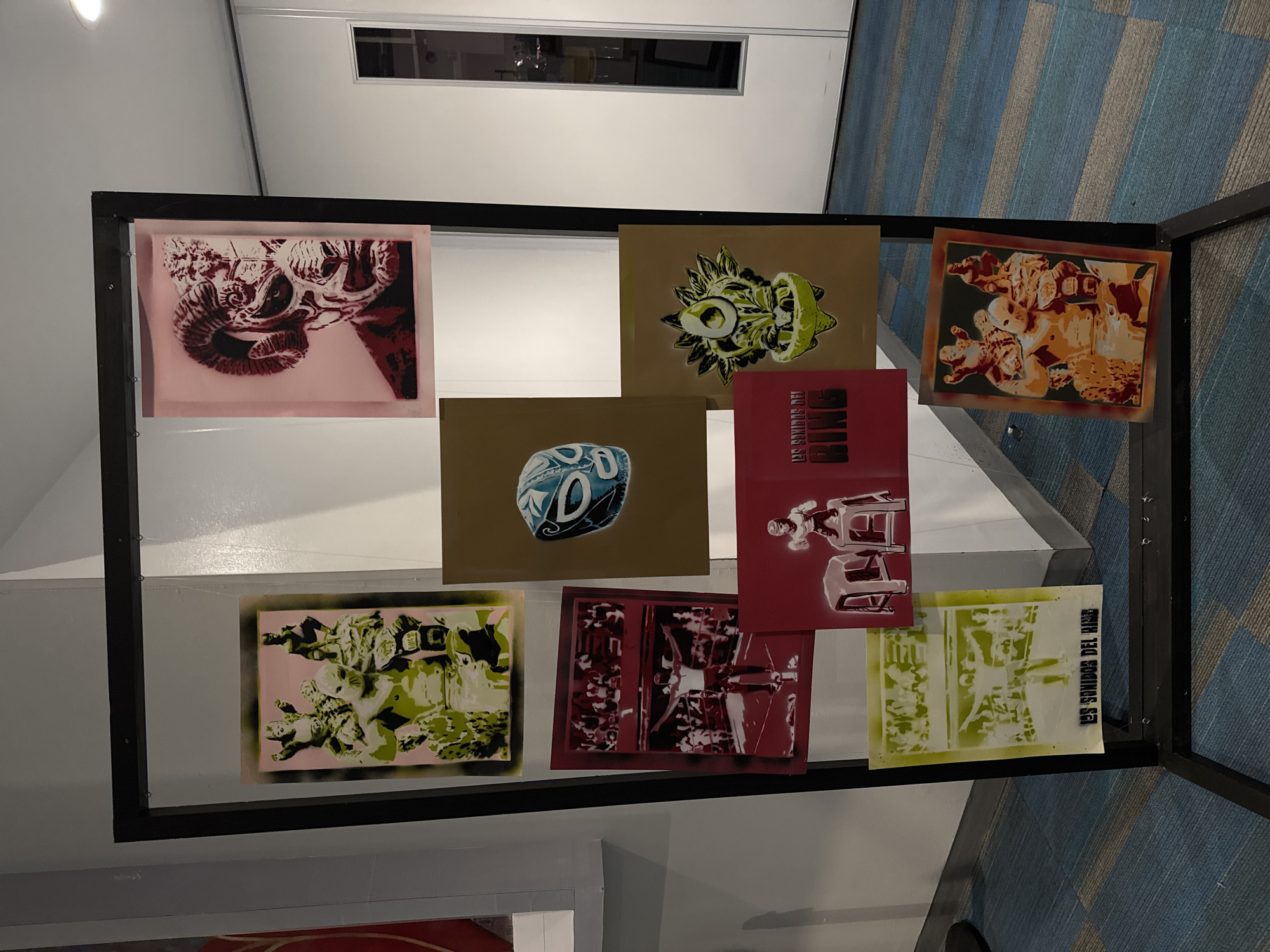 The image depicts an art installation, possibly in an exhibition in Puebla, Mexico. The installation consists of nine framed art pieces arranged in a 3x3 grid within a black metal frame. The art pieces feature diverse themes and color schemes, including imagery related to Mexican wrestling, skulls, and other unidentified subjects. The background features a bright, clean wall and a blue and gray striped floor, suggesting a gallery or exhibition space. The overall impression is of a modern art display featuring cultural and abstract themes.