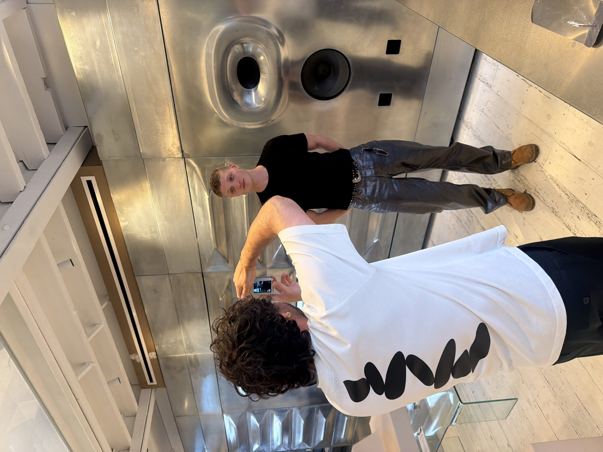 In London, United Kingdom, a man with curly dark hair, wearing a white t-shirt with a black abstract design on the back, photographs another man standing against a large, metallic, textured wall. The man being photographed is wearing a black t-shirt, metallic silver pants, and brown shoes. The metallic wall features two large circular indentations, one above the other, with dark interiors, resembling speaker grilles or decorative elements. Above the metallic wall, a white ceiling with exposed beams is visible, with a horizontal metallic duct or vent system running across it. The floor is made of light-colored wooden planks. A glass table with a metal frame is visible in the background to the left. The overall scene suggests an indoor, possibly retail or gallery, environment.