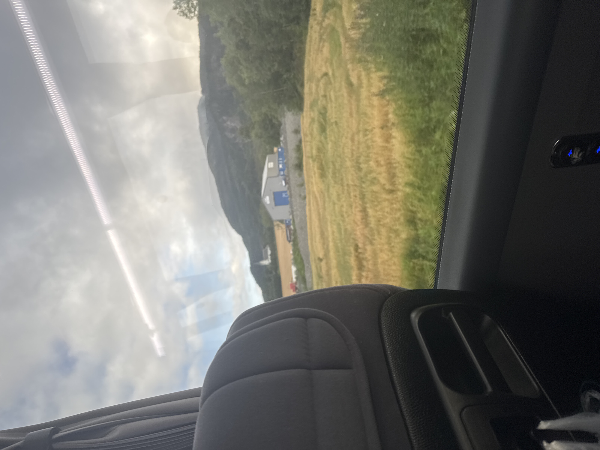 The image is taken from inside a vehicle, looking out the window at a rural landscape. The foreground shows the interior of the vehicle, including a gray, quilted seatback and a black plastic console with storage compartments. A bright white LED light strip is visible across the top of the window, reflecting on the glass.

Outside the window, a field of golden wheat or barley stretches towards rolling hills. In the middle ground, there is a large, modern industrial building with a blue door, situated next to a smaller red structure. Further in the distance, a white church or chapel with a steeple is visible on a hillside. Lush green trees and vegetation cover the hillsides. The sky is partly cloudy, with the sun casting a bright light.

The location is identified as Skatval, Norway. The scene depicts a typical view from a vehicle traveling through the Norwegian countryside, with agricultural fields, industrial buildings, and natural scenery. There are no people visible in the image. The image captures a moment of travel, offering a glimpse of the landscape outside the vehicle.