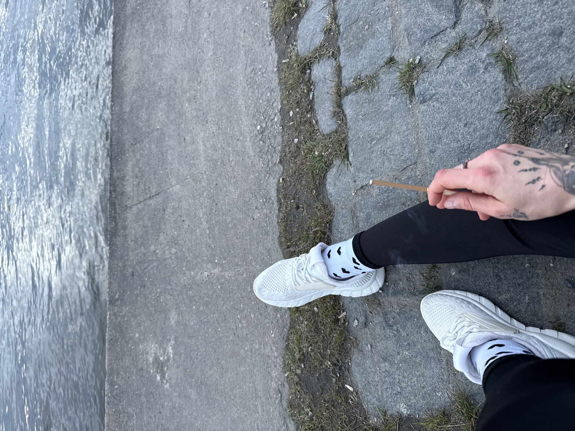 The image captures a high-angle view of a person's lower body, positioned outdoors beside a body of water. The subject is wearing black leggings and white athletic shoes, paired with white ankle socks adorned with scattered black, abstract patterns. One foot is placed closer to the viewer, with the other slightly behind.
A hand, visibly tattooed with multiple small designs on the fingers and a larger, stylized pattern on the wrist/forearm, holds a light brown, hand-rolled cigarette or joint between the thumb and forefinger. A ring is worn on one of the fingers.
The setting appears to be an urban waterfront, likely an embankment or pathway. To the left, a substantial body of water, possibly a river or canal in Saint Petersburg, Russia, shows ripple effects on its surface. To the right, the ground consists of a rough, grey paved surface, possibly a mix of concrete and irregular stone blocks. Patches of dry grass and weeds grow in the cracks and between the paving stones, suggesting a slightly worn or naturalized environment.
The lighting is soft and even, indicative of an overcast day or a time of day with indirect sunlight. No strong shadows are visible. The scene conveys a casual, contemplative moment by the water. No visible text is present in the image.