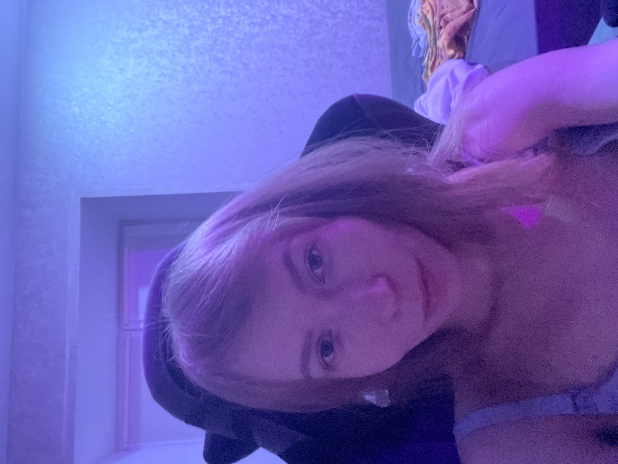 A close-up, low-angle shot shows a woman with long, light-brown hair and fair skin, illuminated by a purple light. She is seated in a black chair. In the background, a white wall with a doorframe is visible. There is a pile of clothing resting on a bed, and the wall is white. The location cannot be determined.