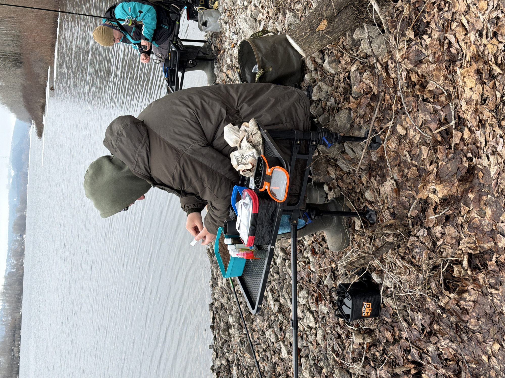 The image depicts two individuals fishing on the bank of a body of water. The foreground shows a person wearing a dark green hooded jacket, olive-colored rubber boots, and seated in a black fishing chair with various fishing accessories, including a blue bait box, small bottles, and orange-framed glasses. The person is focused on preparing bait or tackle. Beside them is a black bag. The background contains a second person wearing a blue and black jacket and a tan hat and also seated in a fishing chair. Both individuals appear to be fishing on the bank of the Votkinsk Reservoir, near the city of Chaikovsky, Perm Krai, Russia. The bank is covered in rocks, dead leaves, and branches. The water is calm and gray, and the opposite bank is visible in the distance.