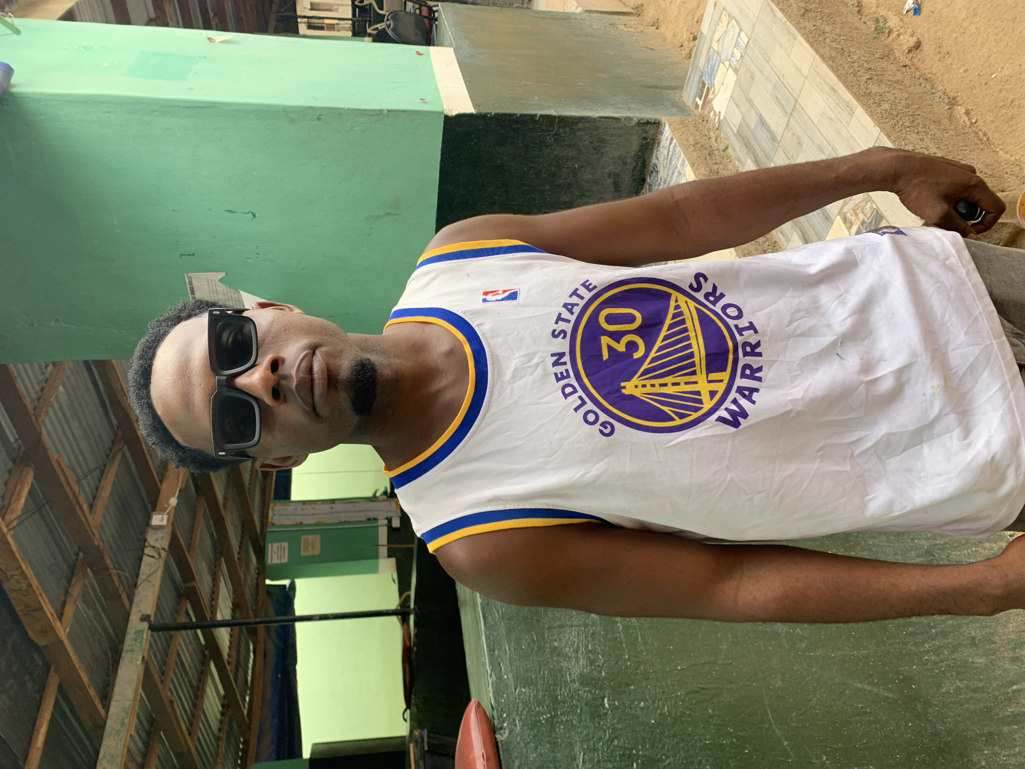 A man stands outdoors, posing for a photograph. He is a young to middle-aged individual with dark skin, short dark hair, and is wearing black rectangular sunglasses. He is dressed in a white sleeveless jersey adorned with blue and yellow trim around the neck and arms. The jersey prominently displays the "Golden State Warriors" logo in purple and yellow, featuring the number "30" within a bridge design. A small NBA logo is also visible on the front of the jersey. His right hand, partially visible, appears to be holding a small container or object.
The background suggests a semi-outdoor or utilitarian setting. To the man's left, there is a light green painted concrete wall or structure. The ground beneath him is a mix of rough dirt or sand, with some areas featuring patterned tiling. Above and behind him, a corrugated metal roof structure supported by wooden beams is visible. The bright, even lighting indicates it is daytime. The man looks directly at the camera with a neutral and composed expression, partially obscured by his sunglasses. This scene likely takes place in a casual area, possibly near the New Stadium in Nigeria, given the architectural and environmental cues.